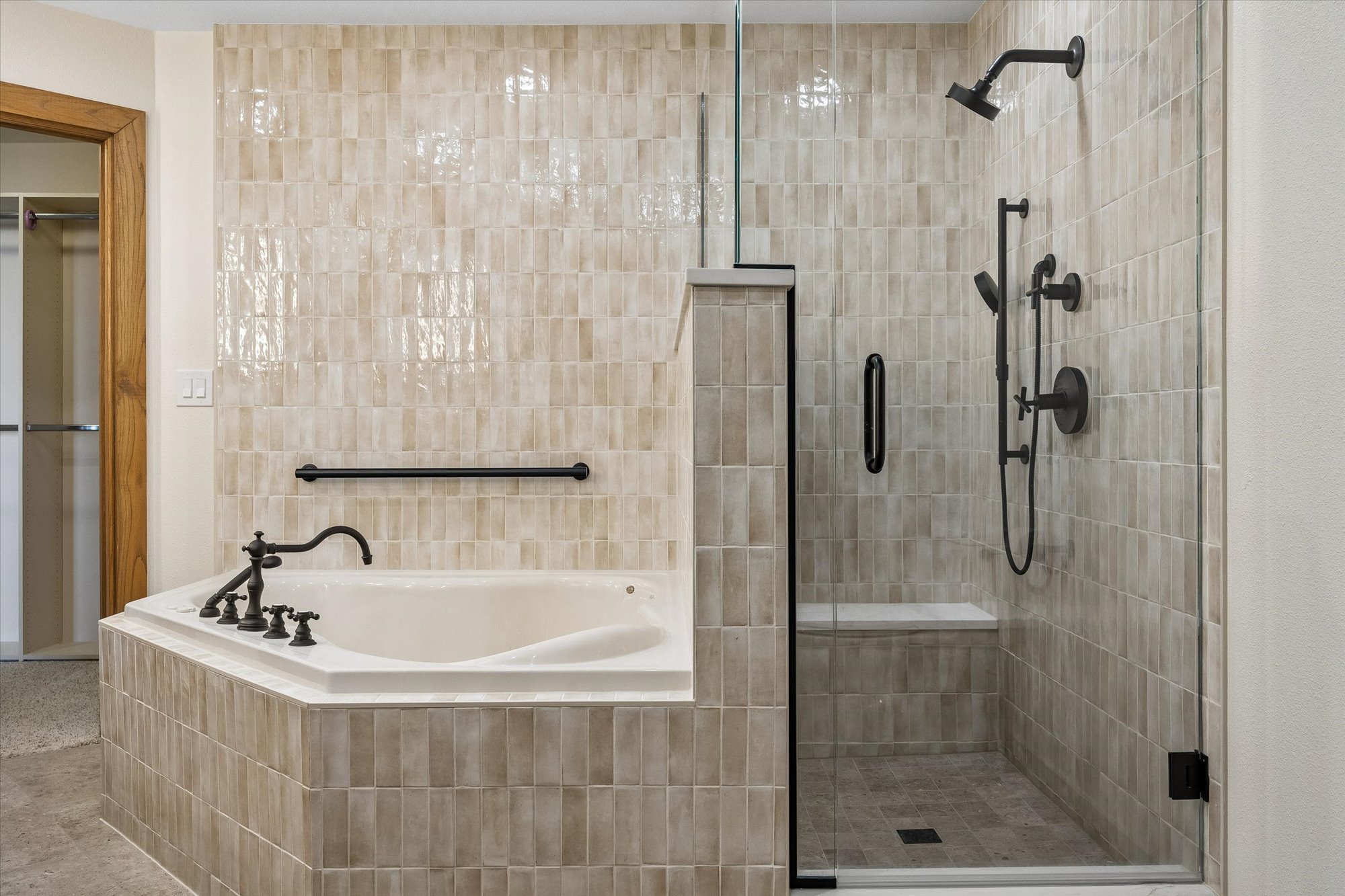 This primary bathroom features a luxurious combination of a soaking tub and a walk-in shower, both covered in elegant, vertical-set beige subway tiles. Matte black fixtures, including a rainfall showerhead and handheld sprayer, provide a modern contrast to the neutral palette, while a glass-enclosed shower stall adds a spacious, open feel. The integration of a built-in shower bench and a safety grab bar above the tub emphasizes both style and accessibility.