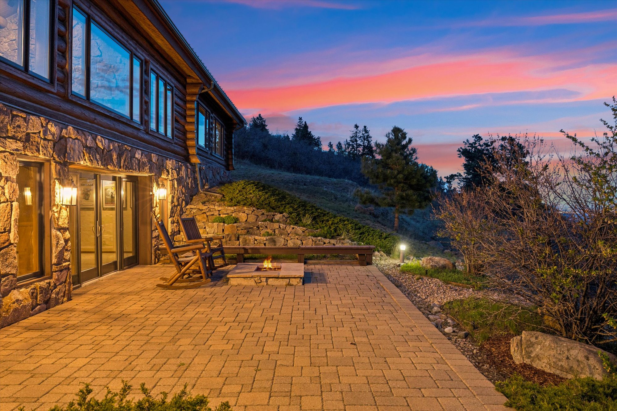 This stunning outdoor patio captures a picturesque sunset view, featuring a stone-clad exterior and a cozy fire pit area with wooden rocking chairs. The paved stone terrace seamlessly blends with the natural landscape, creating an inviting space for evening relaxation or entertaining. The rustic cabin-style architecture and mountain-inspired setting convey a warm, tranquil, and high-end living environment.