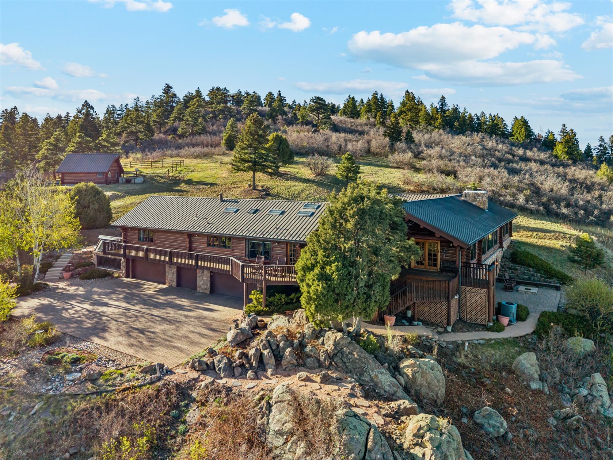 This stunning aerial view showcases a sprawling rustic log cabin home nestled into a scenic, rocky hillside landscape under a bright blue sky. The property features a spacious multi-level wooden deck, a multi-car garage, and a detached outbuilding, all surrounded by dense evergreens and natural terrain. It conveys a tranquil, secluded mountain retreat aesthetic that is both charming and well-integrated into its rugged environment.