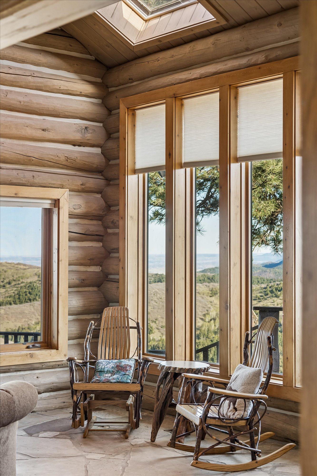 This cozy, rustic living space features log-style wood walls, stone flooring, and expansive windows that offer breathtaking views of the surrounding mountains. The room is furnished with two traditional rocking chairs and a matching side table, creating a perfect nook for relaxation. A skylight overhead enhances the natural light, contributing to the warm and inviting mountain lodge aesthetic.