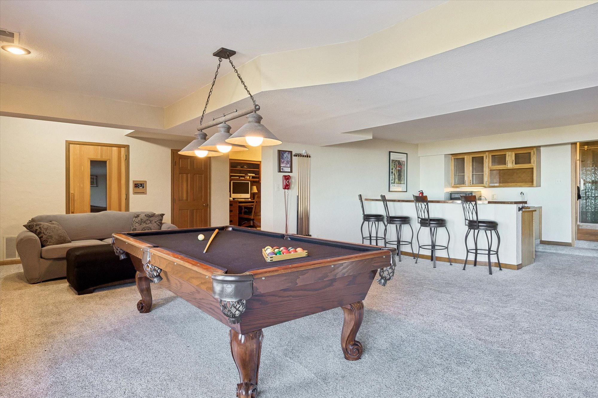 This finished basement functions as an expansive recreation area, featuring a prominent billiard table as its central focal point. The space includes a comfortable seating area with a sofa, a built-in wet bar with high-top seating, and a separate nook designated as an office space. The neutral wall colors and wall-to-wall carpeting create a cozy, multi-purpose environment ideal for entertainment and relaxation.
