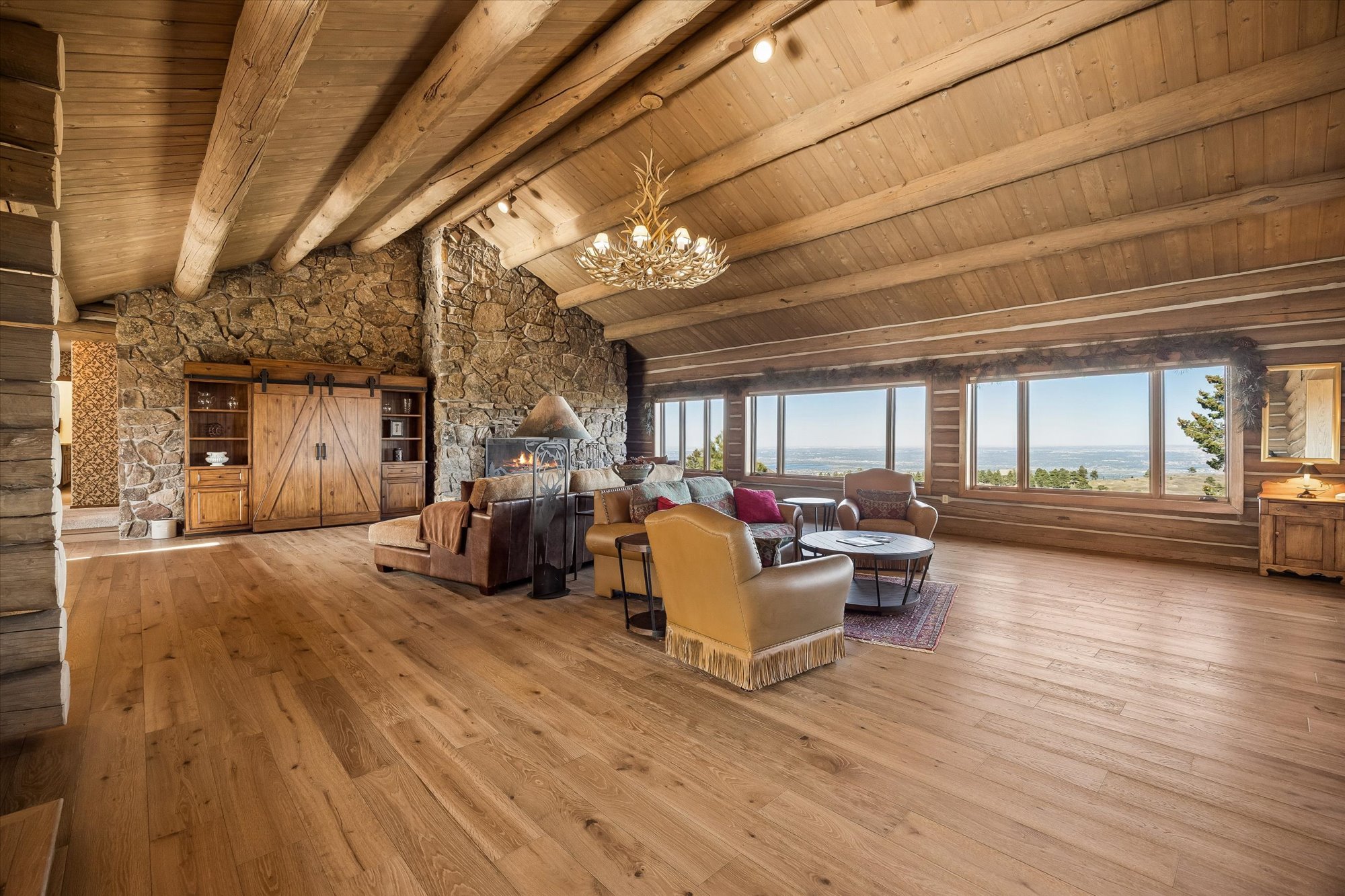 This spacious, rustic-style living room features exposed timber ceiling beams, a striking floor-to-ceiling stone fireplace, and polished wood floors. Large windows provide expansive panoramic views, while the cozy seating area is accented by a distinctive antler chandelier, creating a warm and sophisticated mountain retreat atmosphere.