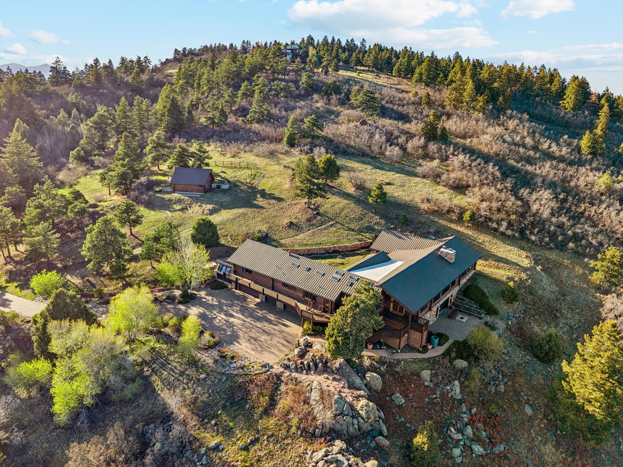 This stunning aerial view captures a sprawling, rustic log home situated on a rugged, hillside landscape dotted with pine trees and rocky terrain. The residence features a durable dark metal roof, multiple expansive decks for outdoor living, and a paved driveway leading to the property, which also includes a separate smaller log cabin nearby. The property offers a serene, elevated mountain aesthetic with a perfect blend of natural beauty and secluded living.
