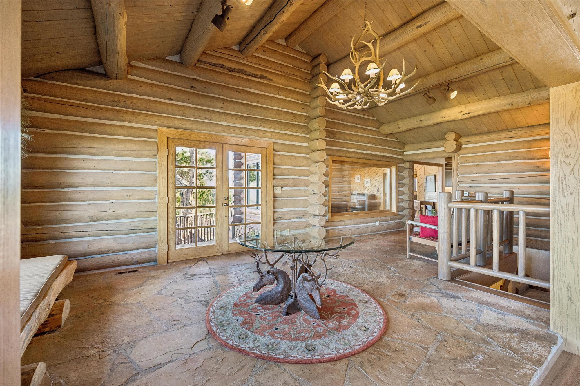 This rustic entryway or living area features high vaulted ceilings with exposed log beams and a grand antler chandelier. The space showcases a unique glass-topped table supported by deer sculptures, complemented by natural stone flooring and warm, horizontal log walls. The presence of a doorway leading to an outdoor deck and an open staircase suggests an inviting, cabin-style dwelling with high-end, thematic mountain decor.