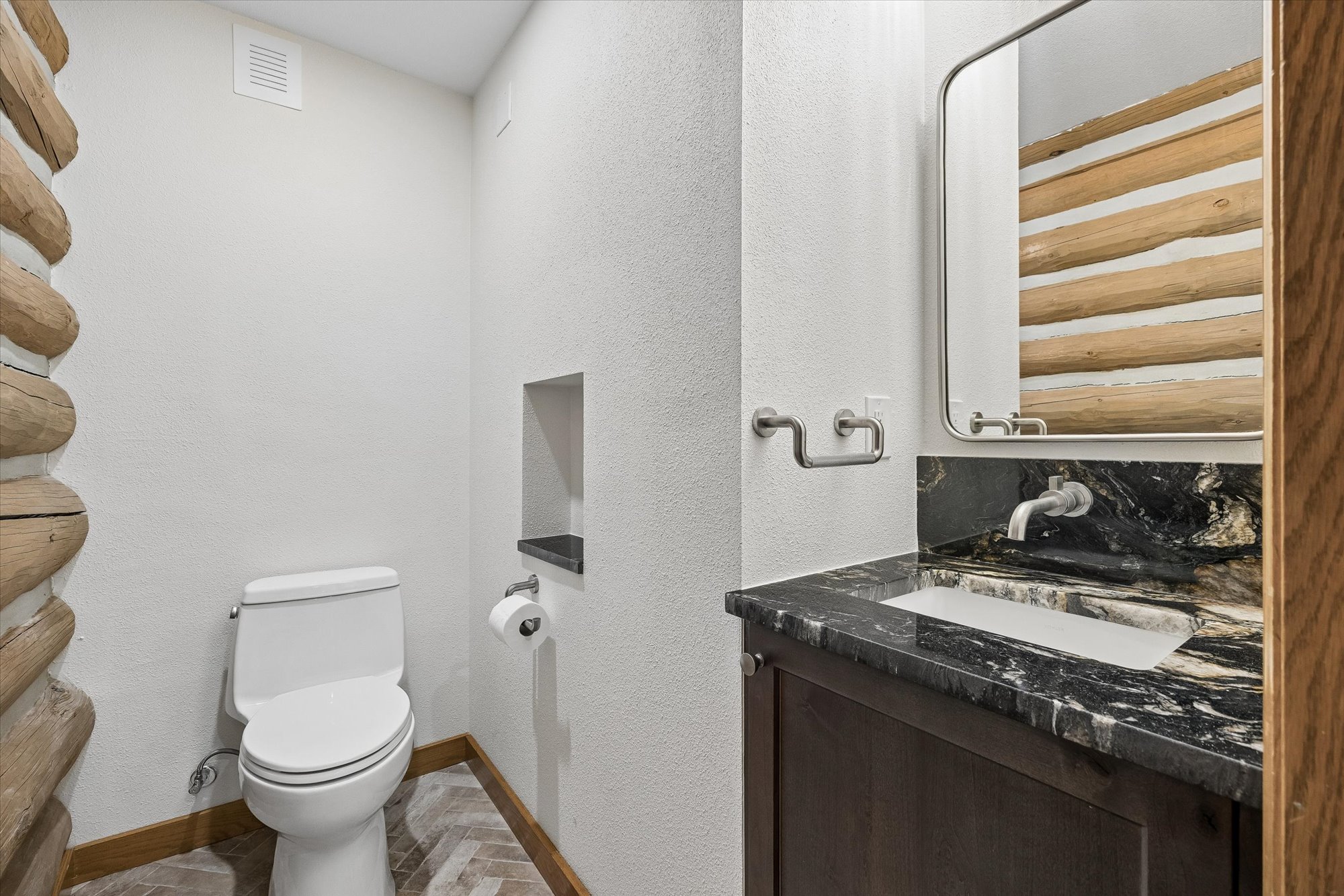 This rustic powder room features a distinctive log-wall accent that transitions into white textured walls, blending cabin-style charm with a modern vanity. The vanity is topped with a dramatic black marble countertop and an integrated sink, complemented by stylish wall-mounted hardware and a contemporary mirror. The combination of high-end finishes and rugged natural elements creates a sophisticated yet cozy atmosphere.