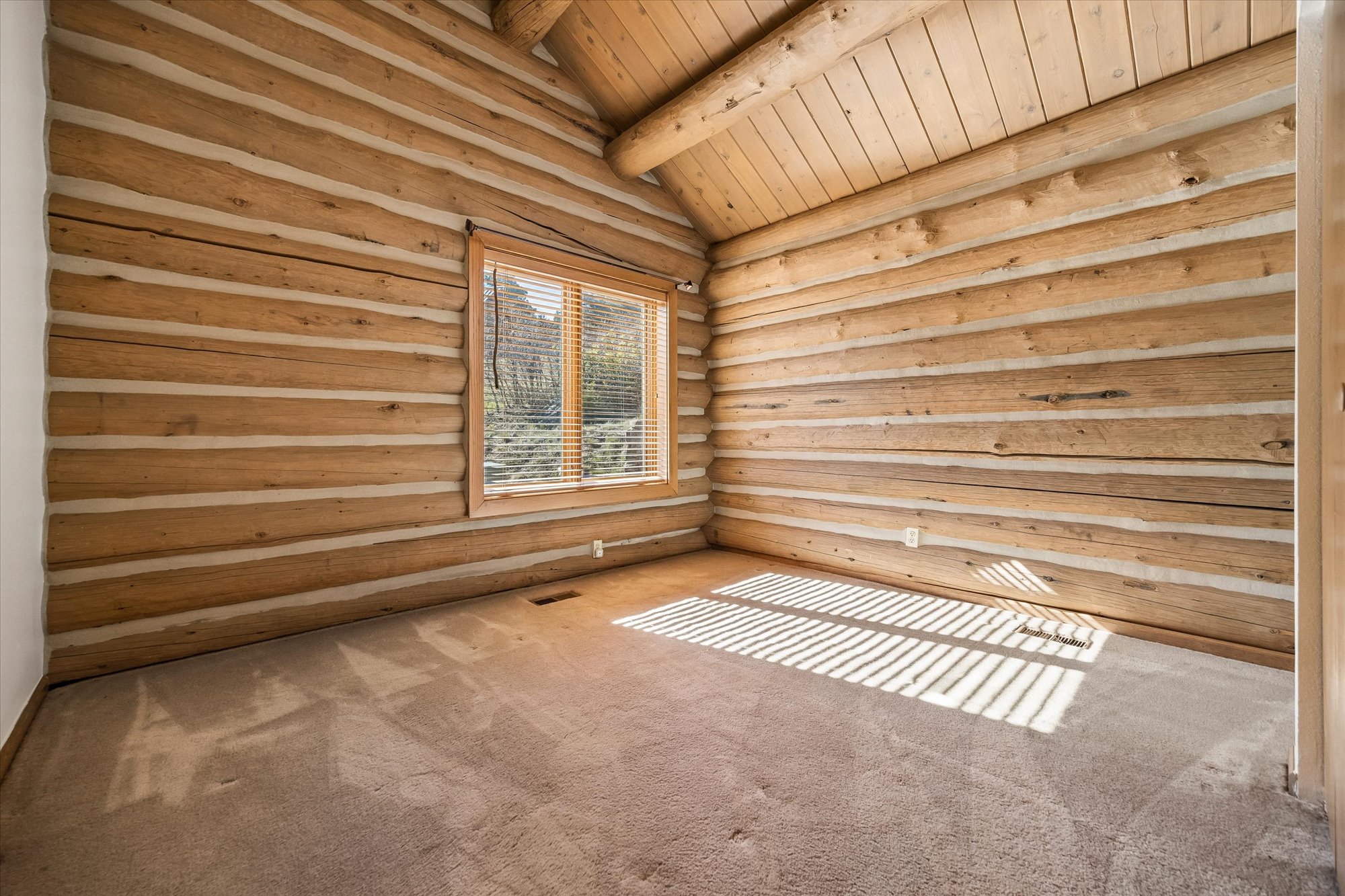 This is a rustic, light-filled bedroom characterized by its exposed log walls and vaulted wood-plank ceiling, which provide a cozy, cabin-like aesthetic. The room is carpeted in a neutral tone and features a single window with blinds that cast striped shadows across the floor. An empty, clean space, it offers a warm and inviting atmosphere suitable for a guest bedroom or versatile living area.