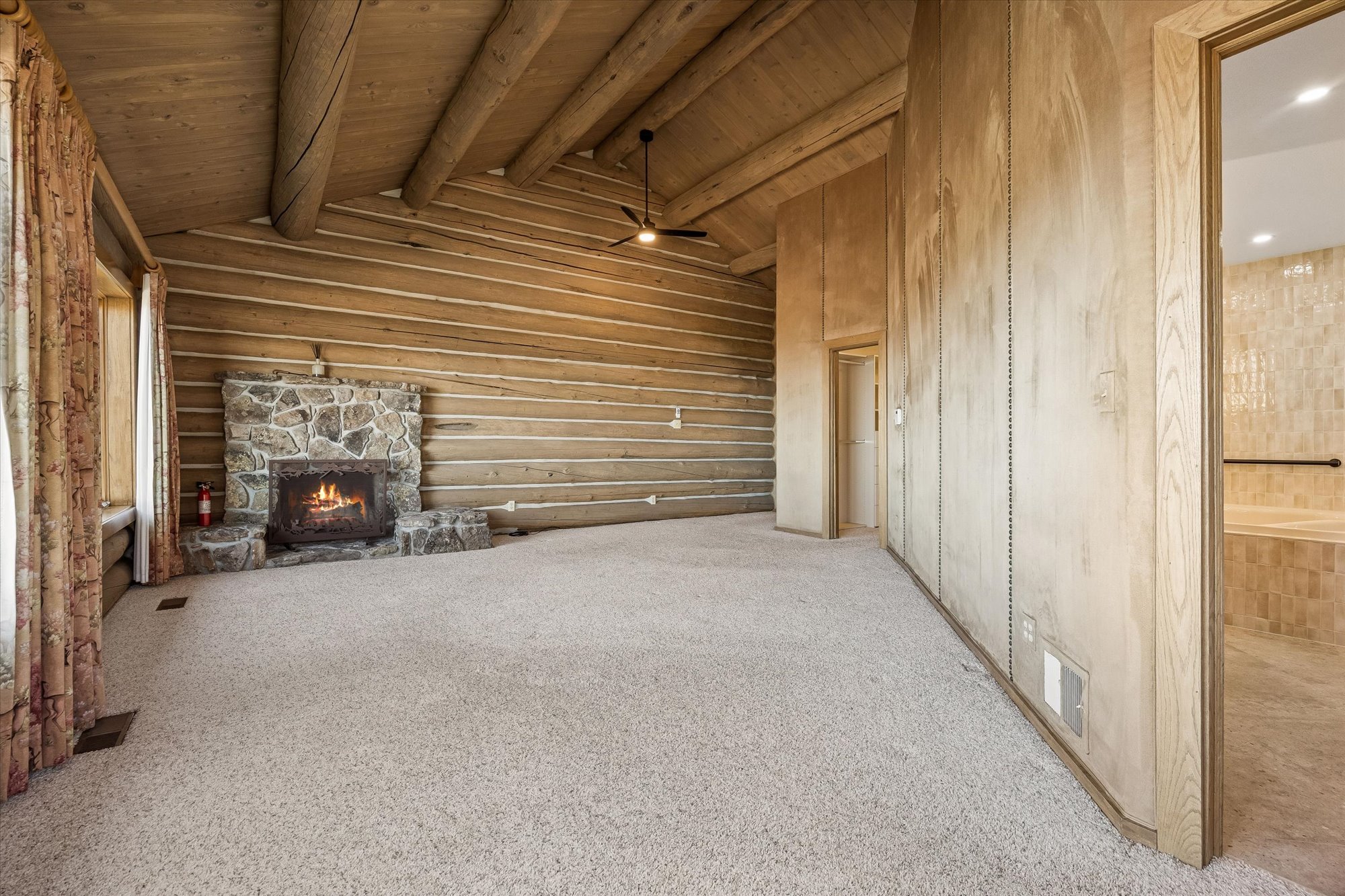 This rustic primary bedroom features full log walls with a vaulted timber-beam ceiling, creating a warm and authentic cabin atmosphere. The room is anchored by a large stone fireplace and features plush neutral-toned carpeting, with an attached bathroom visible through an open doorway. A ceiling fan provides air circulation, and long, decorative curtains frame the window area.