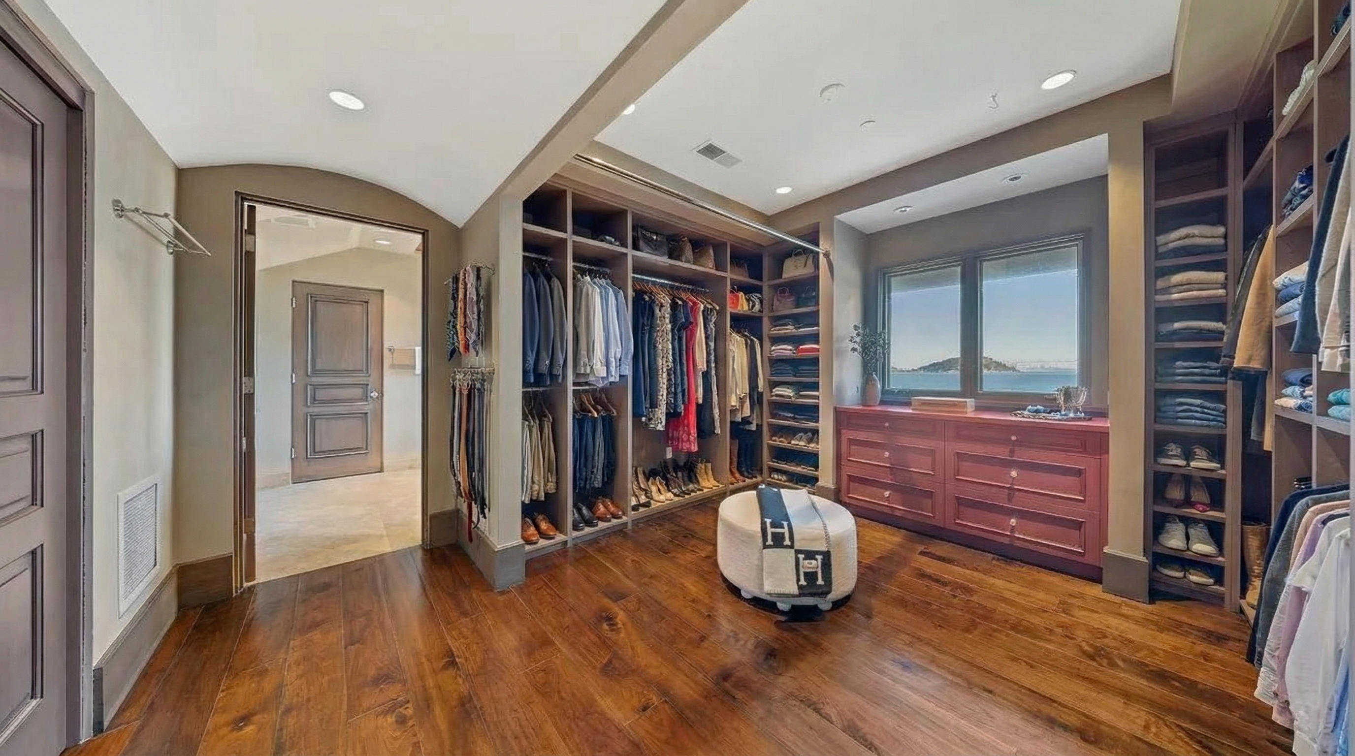 This is a spacious walk-in closet with custom shelving and rods for organized storage. The closet features wood floors, recessed lighting, and a window providing natural light and views. An ottoman is placed in the center of the room, creating a luxurious dressing area.