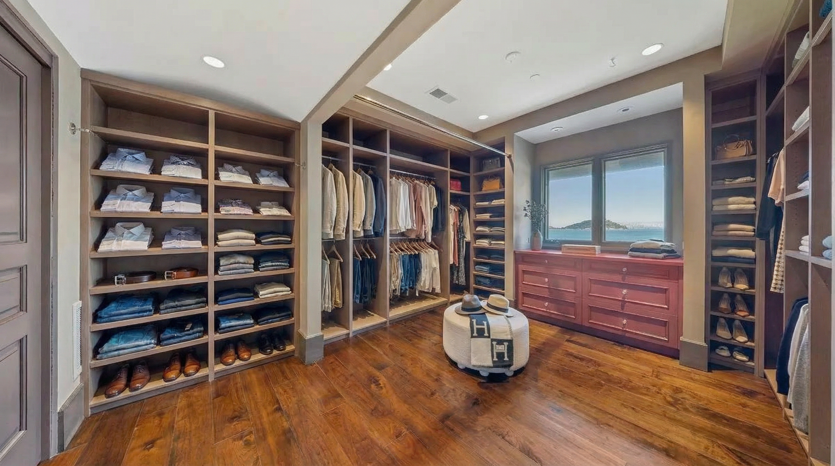 This is a well-organized walk-in closet with custom shelving, hanging rods, and drawer units. The closet features a mix of open and closed storage, showcasing neatly folded clothes, shoes, and accessories. Hardwood floors and recessed lighting create a luxurious and functional space.