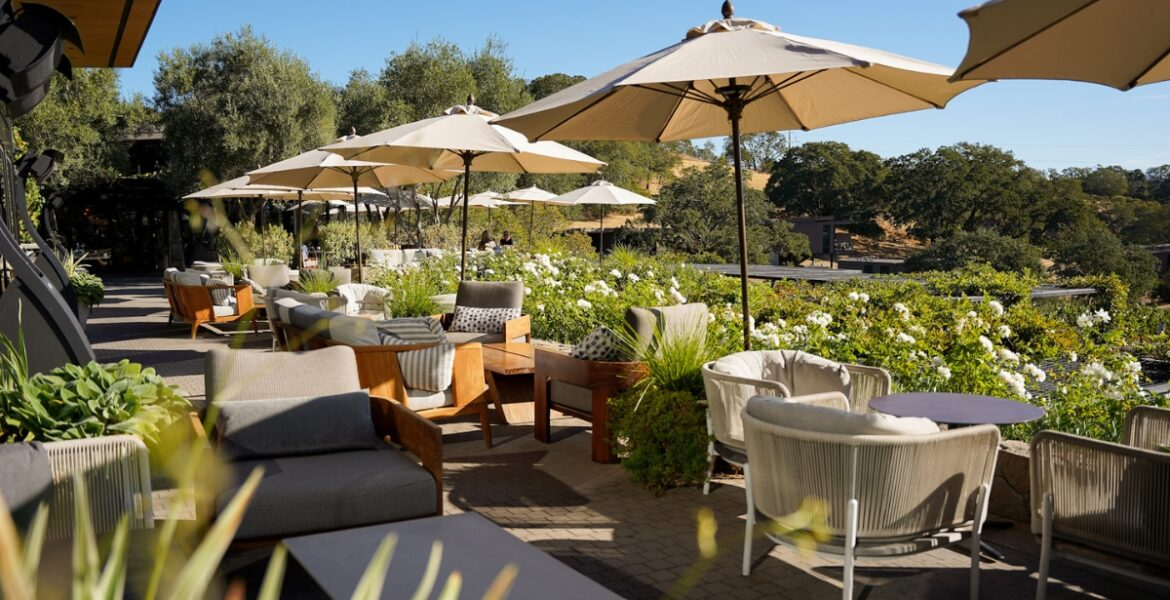 This spacious, open-air terrace is furnished with sophisticated lounge seating, including plush armchairs and sofas arranged for relaxation. The area is shaded by multiple neutral-colored umbrellas and features lush white flowering landscaping, creating a serene, upscale atmosphere. The patio offers a scenic overlook, making it an ideal setting for outdoor dining or entertaining in a luxury residential or hospitality environment.
