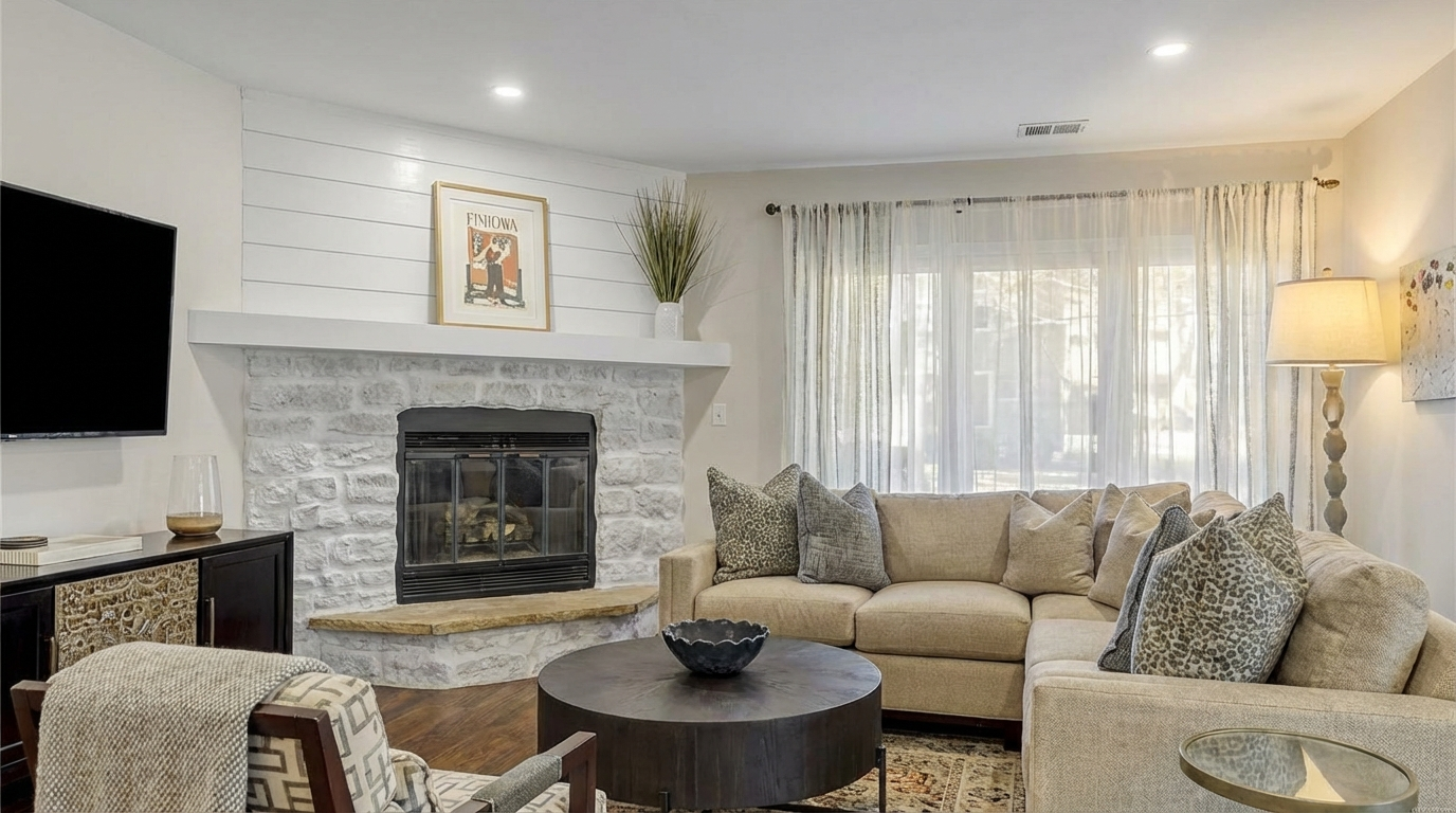 This is a cozy and inviting living room featuring a stone fireplace with a white mantel and shiplap above. A comfortable sectional sofa is arranged around a dark round coffee table. Light-filtering curtains allow natural light to fill the space, creating a bright and airy atmosphere.