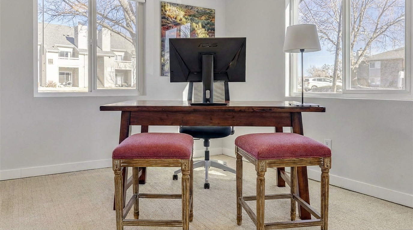 The image showcases a well-lit home office featuring a dark wood desk with an ASUS monitor and a table lamp. Two decorative stools upholstered in red fabric are positioned in front of the desk. Natural light fills the room through two large windows, creating a bright and inviting workspace.