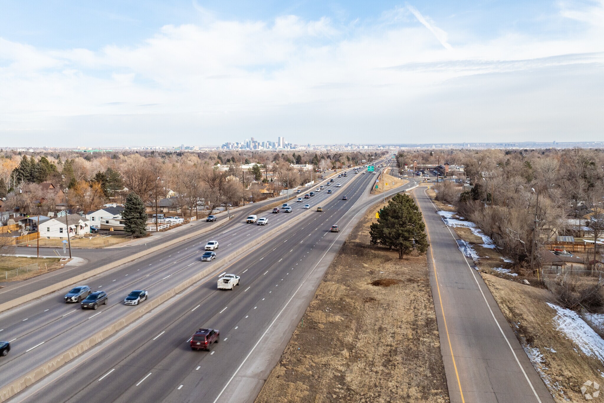 This aerial shot captures a wide view of a highway surrounded by mature trees and residential areas. In the distance, a city skyline is visible, adding an urban element to the suburban landscape. The scene conveys a sense of connectivity and accessibility, highlighting the property's location relative to both nature and the city.