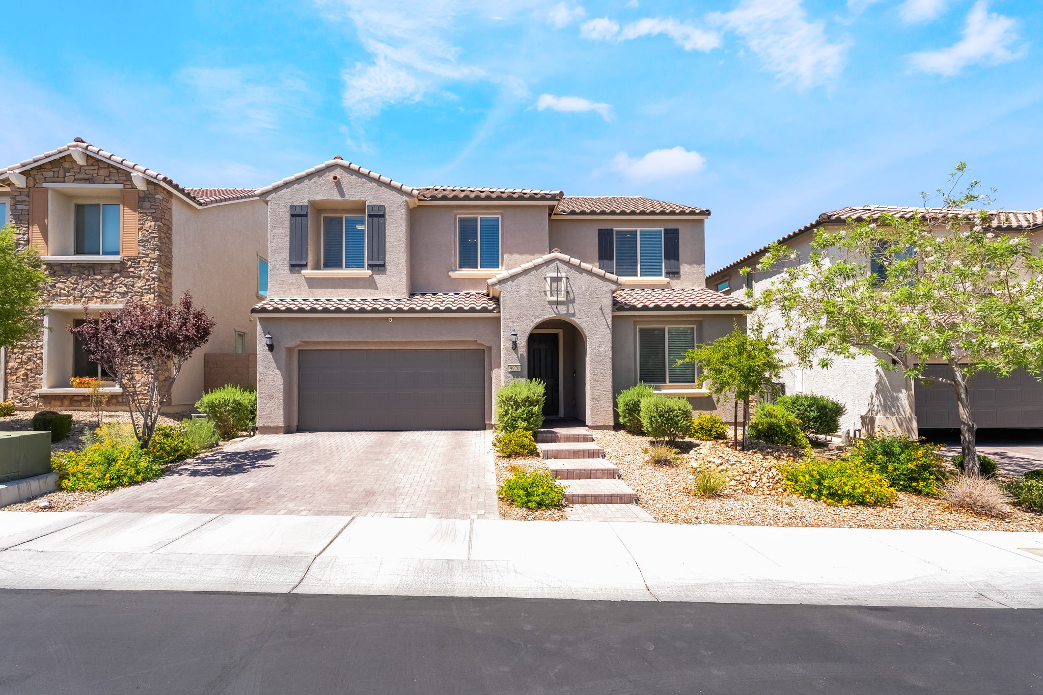 This two-story suburban home features a stucco facade with a tiled roof and a paved driveway leading to a two-car garage. The front entryway is recessed under a covered porch, complemented by desert-style landscaping with shrubs and small trees. The perspective is a straight-on, eye-level shot capturing the home's symmetrical design and curb appeal under a bright blue sky.