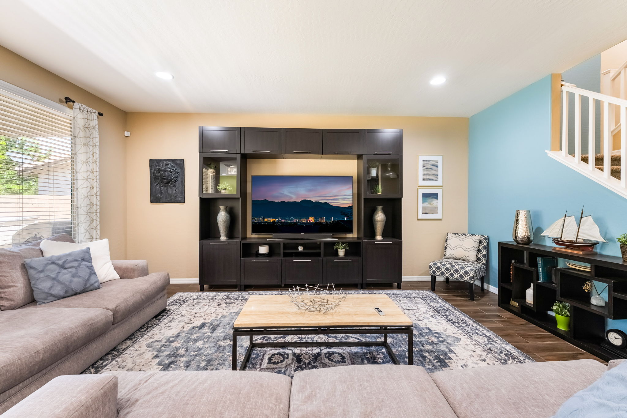This inviting living room features a large, dark wood entertainment center as the focal point, housing a television and decorative accents. A spacious sectional sofa faces the entertainment unit, centered around a light wood coffee table on a patterned area rug. The room is accented by a vibrant blue wall near the staircase and neutral-toned walls, creating a balanced and comfortable living space.