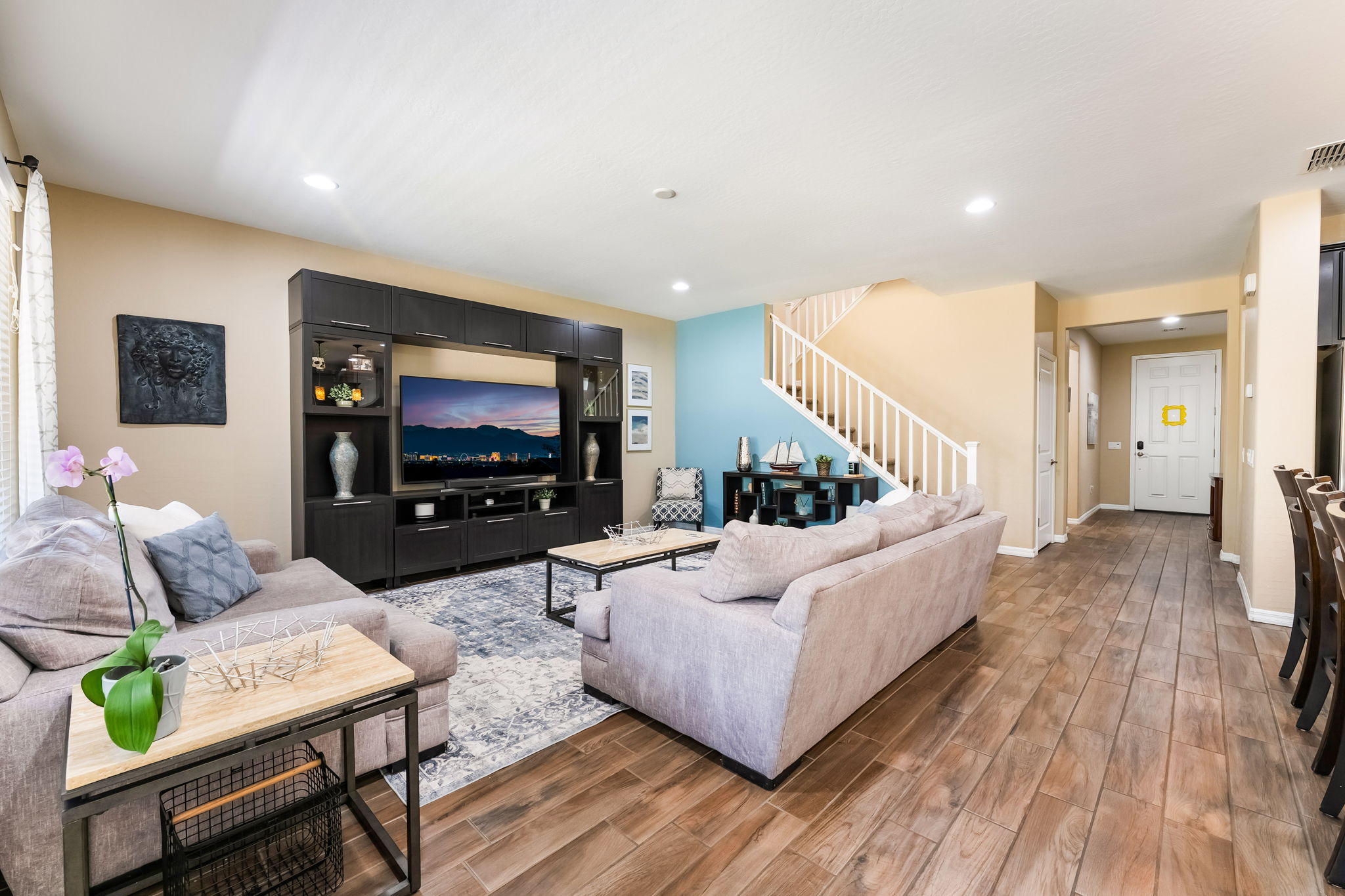 This inviting living room features a large, dark wood entertainment center housing a television, complemented by a light-colored sofa and a patterned area rug. The space is accented by a vibrant blue wall near the staircase, which adds a modern touch to the neutral color palette. Wood-look tile flooring flows throughout the open-concept area, creating a cohesive and warm atmosphere perfect for relaxation.