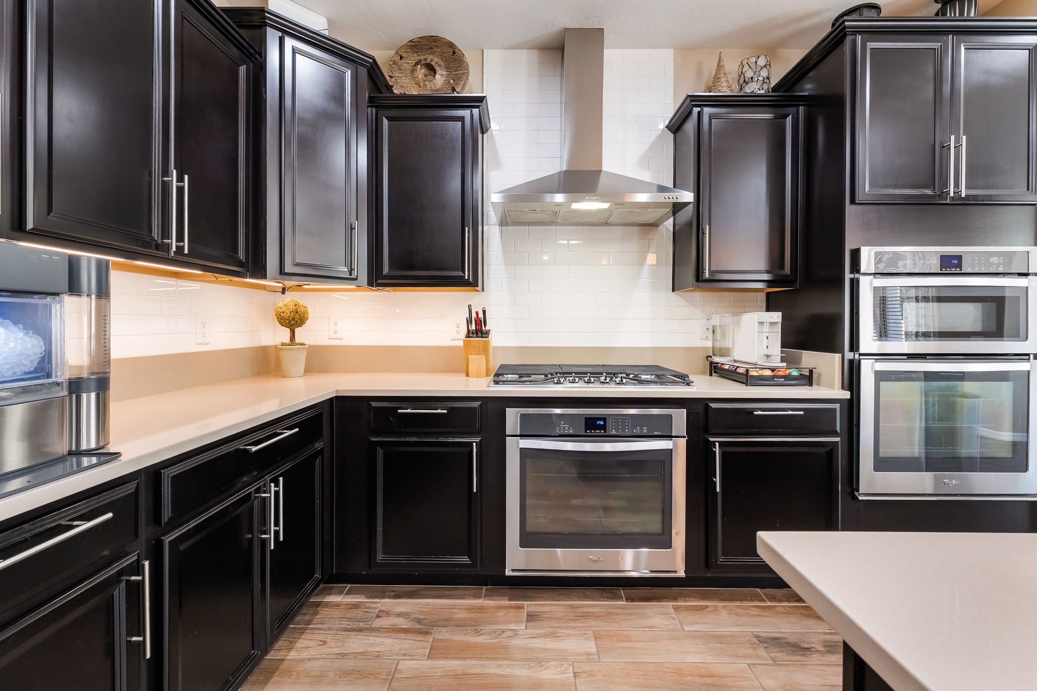 This modern kitchen features dark cabinetry contrasted by light-colored countertops and a white subway tile backsplash. The space is equipped with stainless steel appliances, including a gas cooktop, a built-in oven, and a double wall oven unit. The perspective is a straight-on view of the main cooking wall, highlighting the clean lines and functional layout of the workspace.