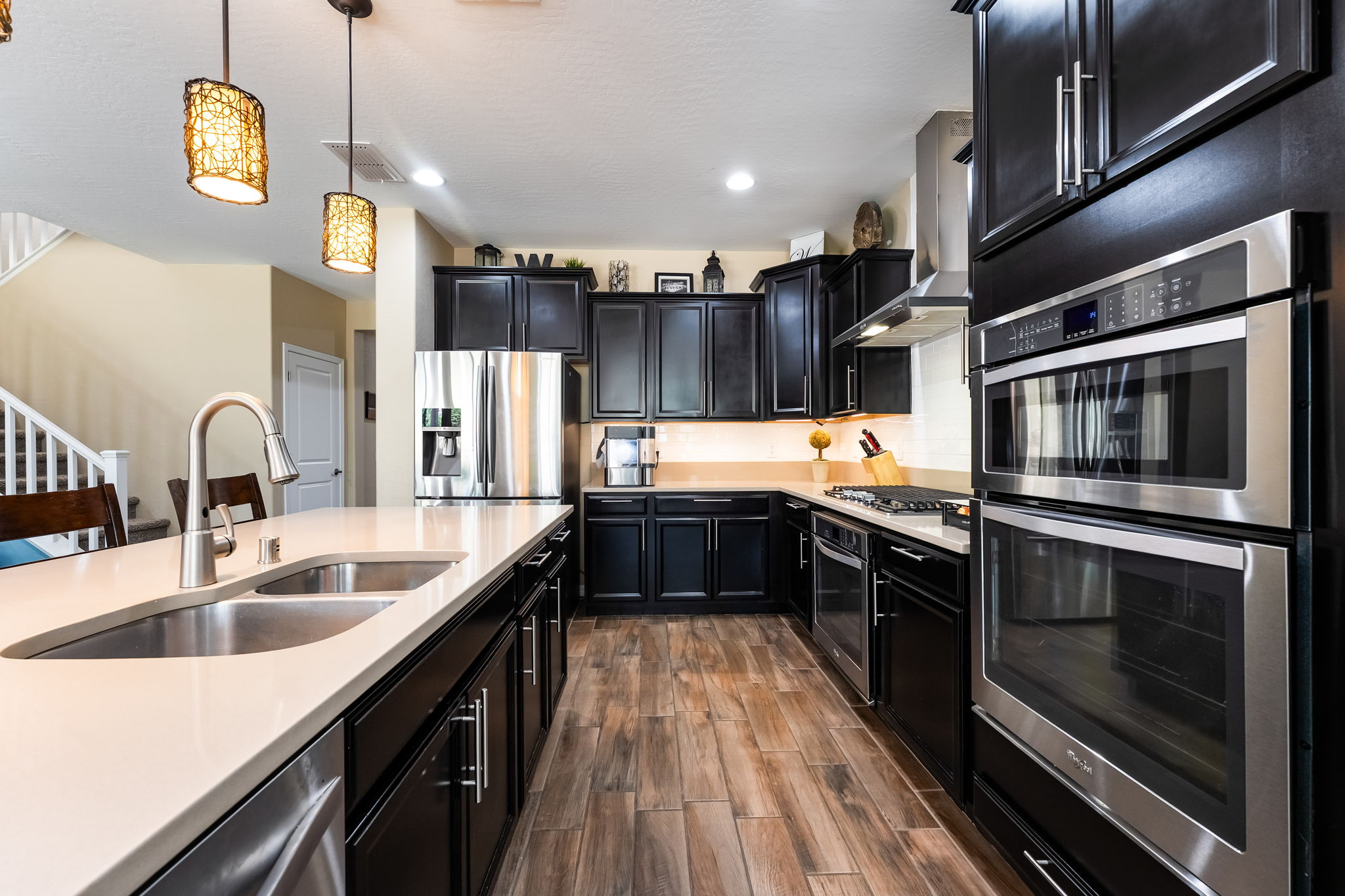 This modern kitchen features dark cabinetry paired with light-toned countertops and stainless steel appliances, including a double wall oven and a side-by-side refrigerator. A large central island with a double sink provides ample workspace, while two decorative pendant lights hang above. The space is finished with wood-look tile flooring and under-cabinet lighting, creating a warm and functional culinary environment.