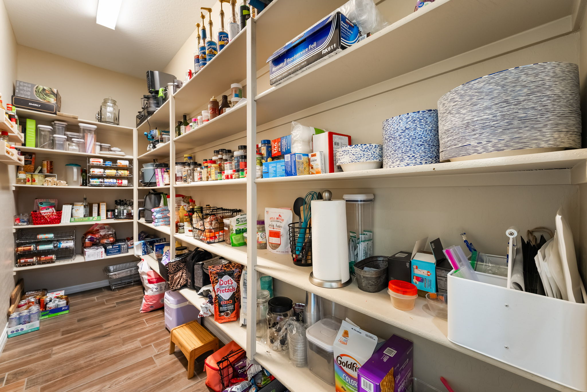 This organized walk-in pantry features multiple levels of white shelving stocked with a variety of food items, paper goods, and kitchen supplies. The space is finished with wood-look tile flooring and a small wooden step stool, creating a functional and well-utilized storage area. The perspective is from the doorway, looking into the depth of the pantry to showcase its ample storage capacity.