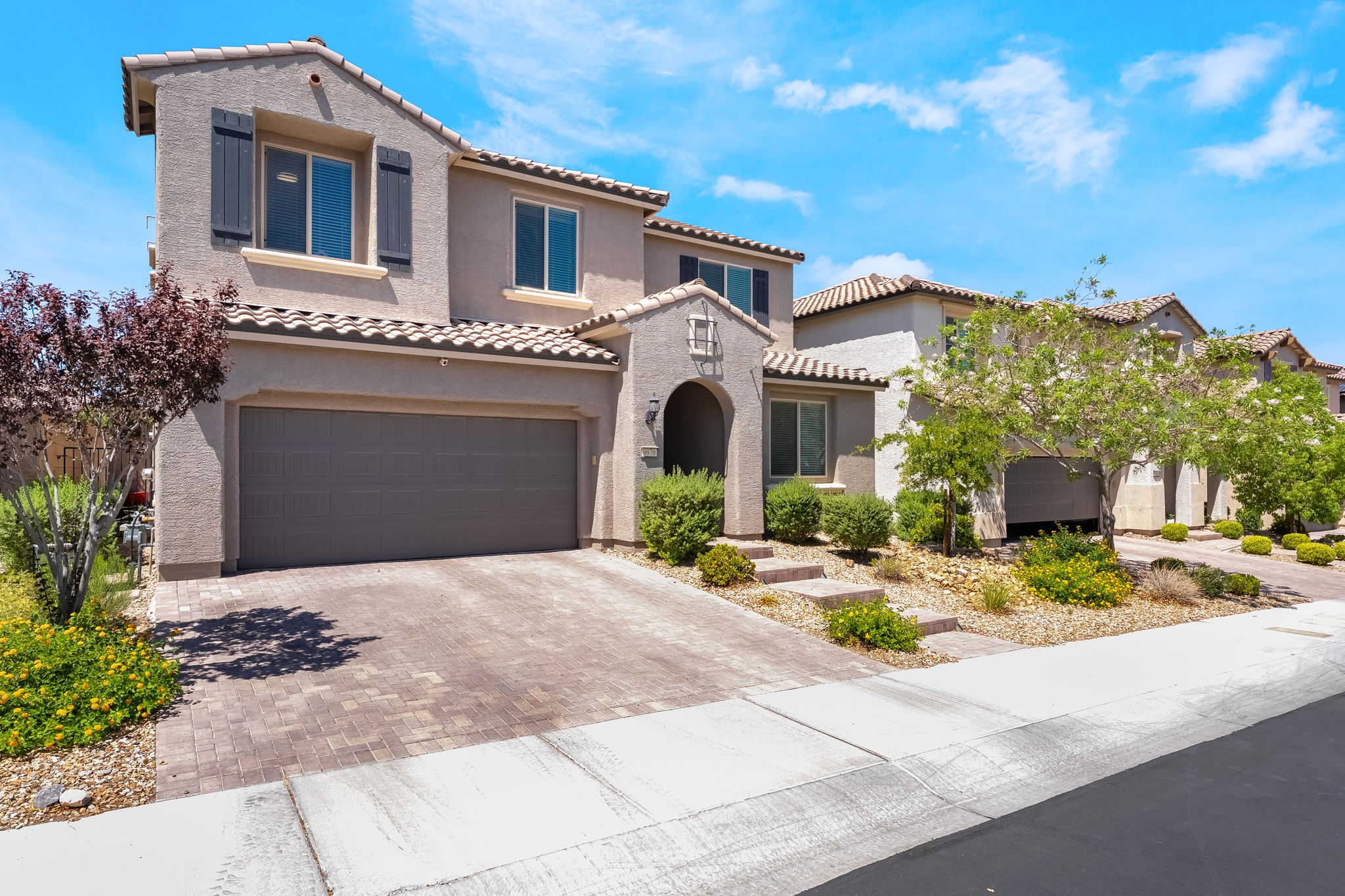 This two-story suburban home features a stucco facade with a tiled roof and a spacious two-car garage. The front yard is landscaped with desert-style plantings, including small shrubs and a mature tree, complemented by a paved driveway and a concrete sidewalk. The perspective is a wide-angle street-level shot, capturing the inviting entryway and the neighboring property under a bright blue sky.