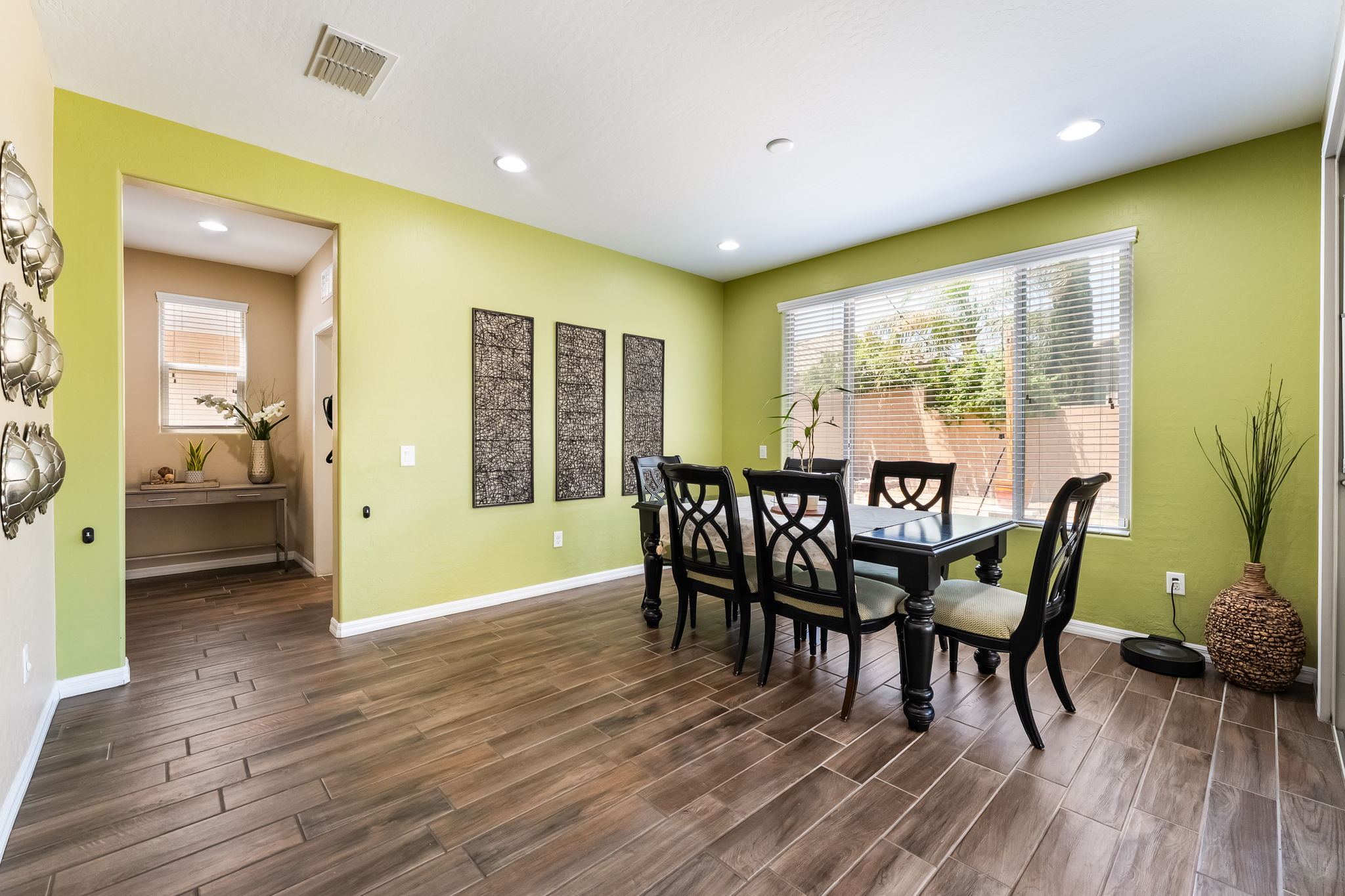 This dining room features a dark wood table with four matching chairs, set against vibrant lime-green walls and wood-look tile flooring. Three decorative metal wall panels provide a focal point, while a large window with blinds allows natural light to fill the space. A doorway leads to an adjacent room, creating an open and connected feel throughout the home.