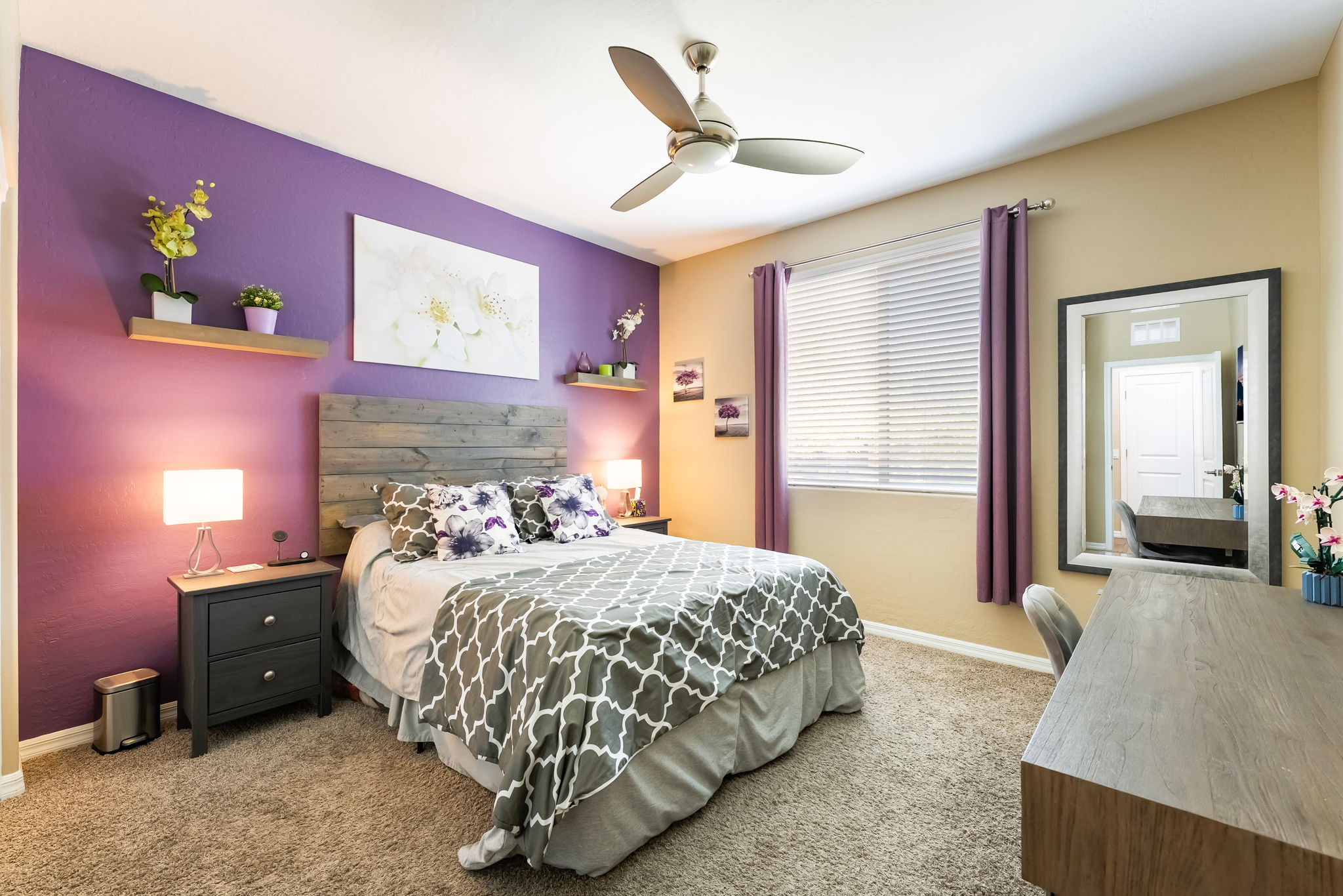 This bedroom features a bold purple accent wall behind a rustic wood-plank headboard, creating a vibrant focal point. The room is furnished with a bed dressed in a grey and white patterned comforter, a dark wood nightstand, and a matching desk area with a large wall mirror. A ceiling fan provides air circulation, while natural light filters through blinds on the window, which is framed by purple curtains.