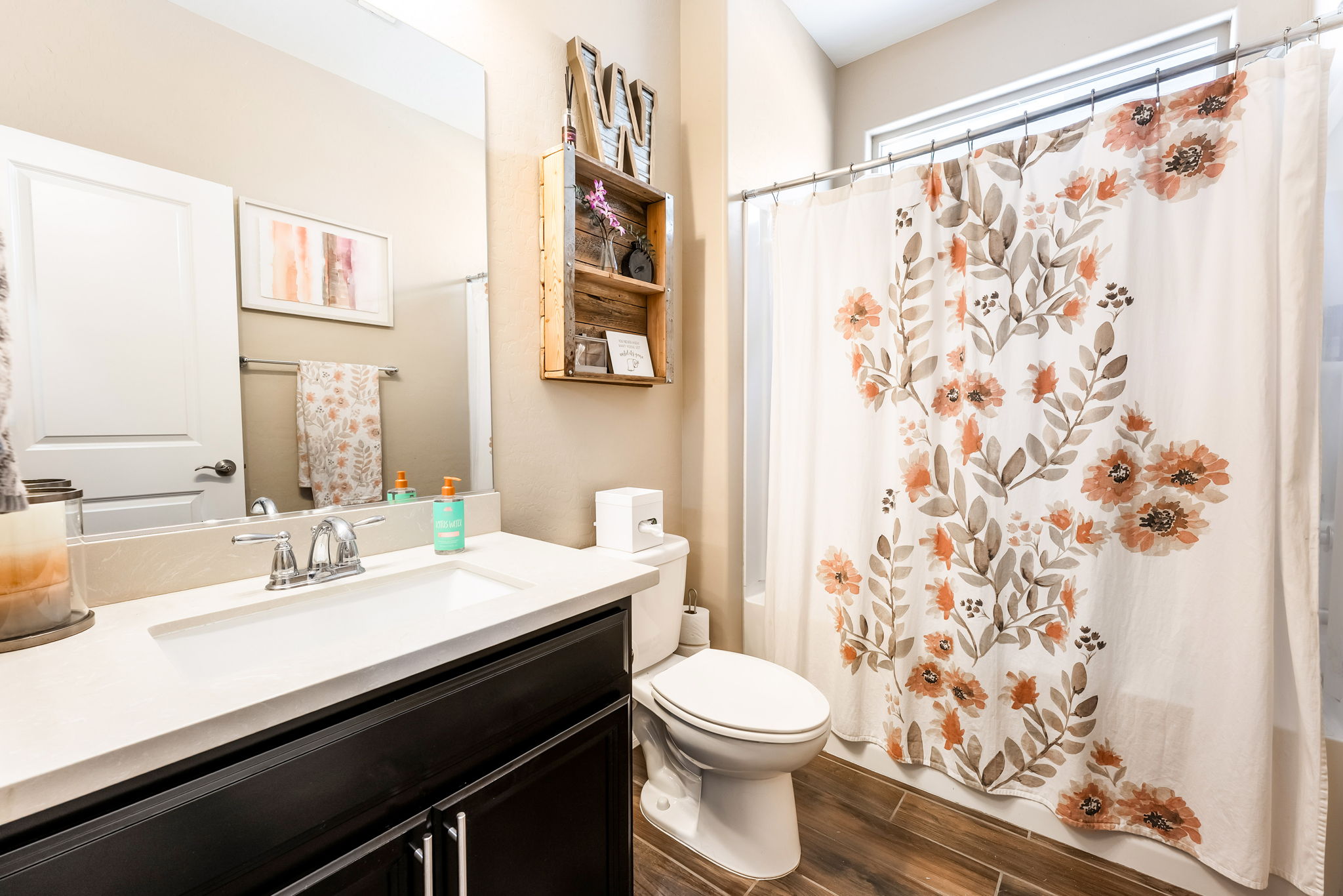 This bright and functional bathroom features a dark wood vanity topped with a light-colored countertop and a chrome faucet. A floral-patterned shower curtain adds a touch of color, while a wooden wall-mounted shelf provides decorative storage above the toilet. The space is finished with wood-look flooring and a large mirror, creating a clean and inviting atmosphere.