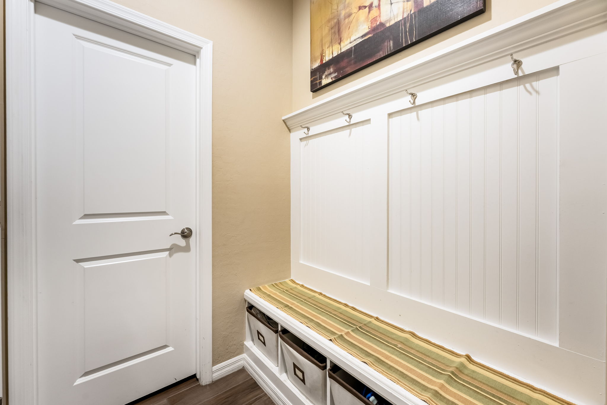 This entryway mudroom area features a built-in white bench with integrated storage cubbies and a matching beadboard wall paneling system equipped with coat hooks. A striped runner adds a touch of color to the bench, while a piece of abstract art hangs above the hooks, creating a functional and organized transition space. The perspective is a straight-on view of the mudroom bench adjacent to a closed white interior door.