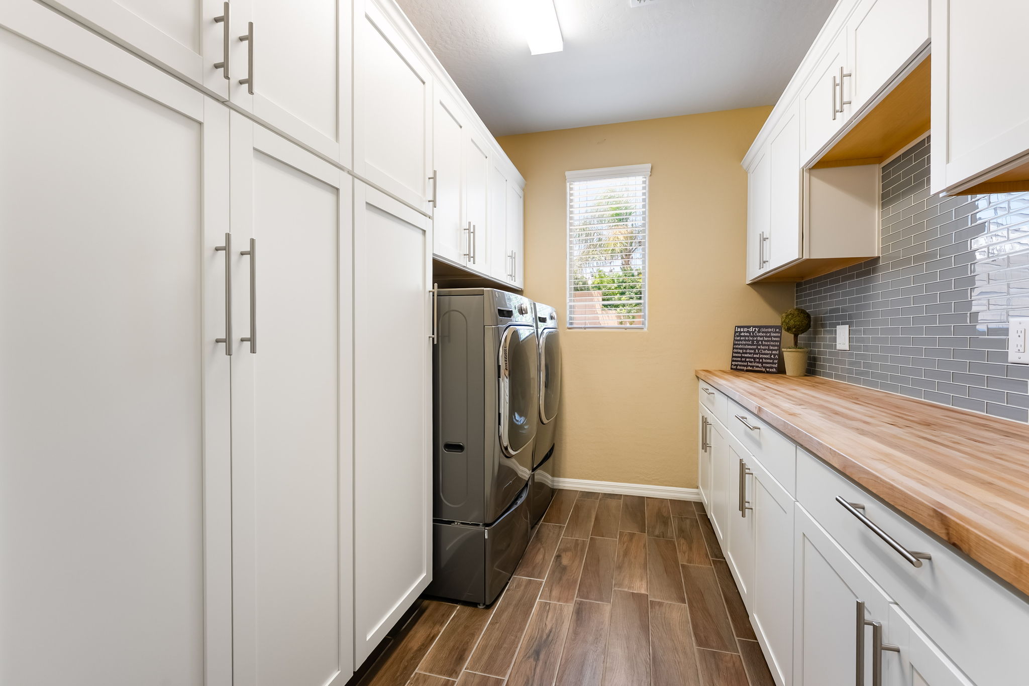 This bright and functional laundry room features crisp white cabinetry providing ample storage, paired with a warm-toned wood countertop and a gray subway tile backsplash. The space is equipped with a modern gray washer and dryer set, complemented by wood-look tile flooring and a window that allows natural light to fill the room. The overall aesthetic is clean, organized, and contemporary, making it a highly practical utility space.