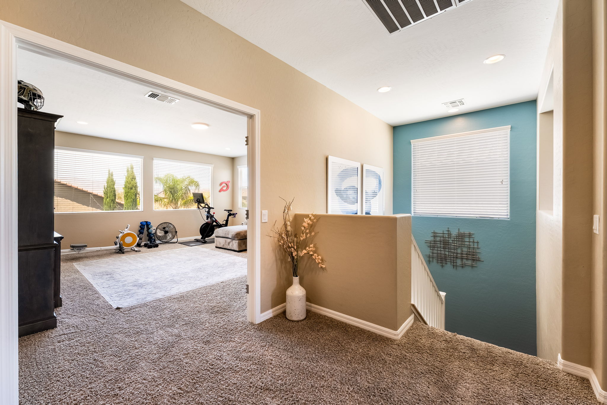 This view from a carpeted landing looks into a home gym through a wide doorway, while a staircase descends to the right alongside a striking teal accent wall. The space features neutral-toned walls, recessed lighting, and a decorative vase with dried branches, creating a clean and functional transition area. The perspective highlights the open floor plan and the connection between the upper-level living space and the lower floor.