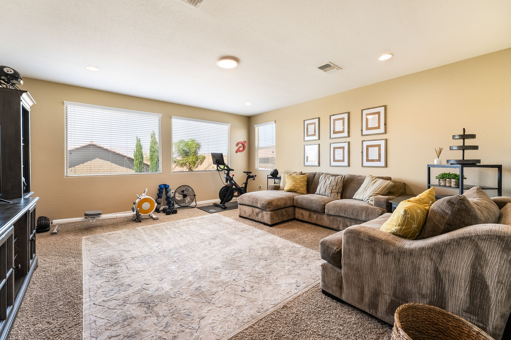 This spacious living room features a large, comfortable sectional sofa arranged around a patterned area rug, creating a cozy seating area. The room doubles as a home gym, with a stationary bike and rowing machine positioned near the windows, which allow for plenty of natural light. The walls are painted a neutral tone and decorated with a grid of framed art, while a dark media console sits to the left, completing the multi-functional layout.