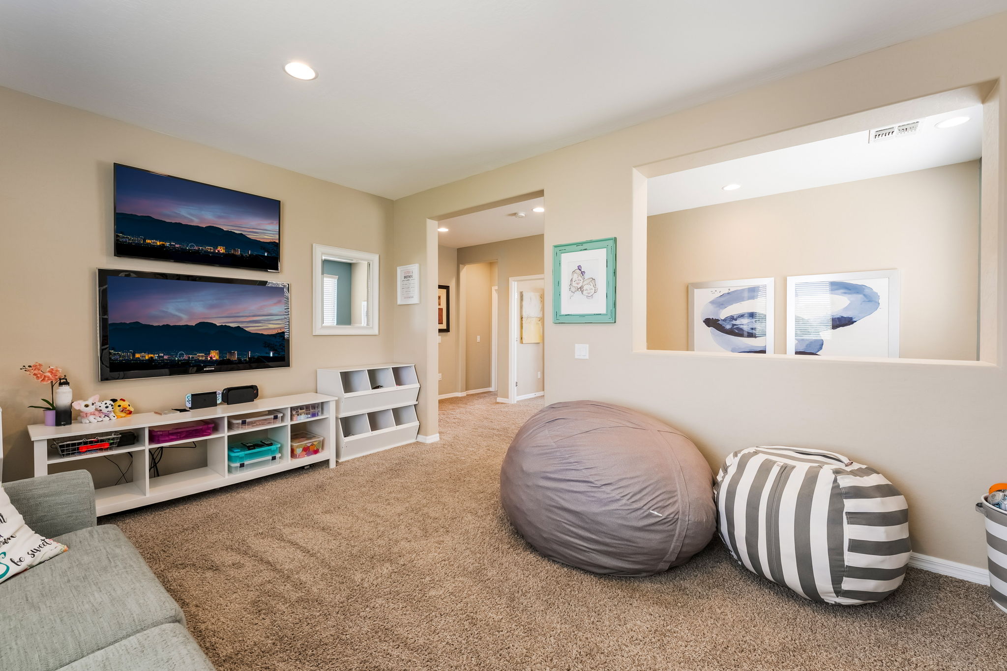 This cozy living room features a neutral color palette with plush carpeting and a comfortable seating area. The space includes a media wall with two stacked televisions, a white storage console, and casual beanbag seating, making it an ideal area for relaxation or entertainment. A large wall cutout provides an open view into an adjacent room, enhancing the sense of space and connectivity throughout the home.