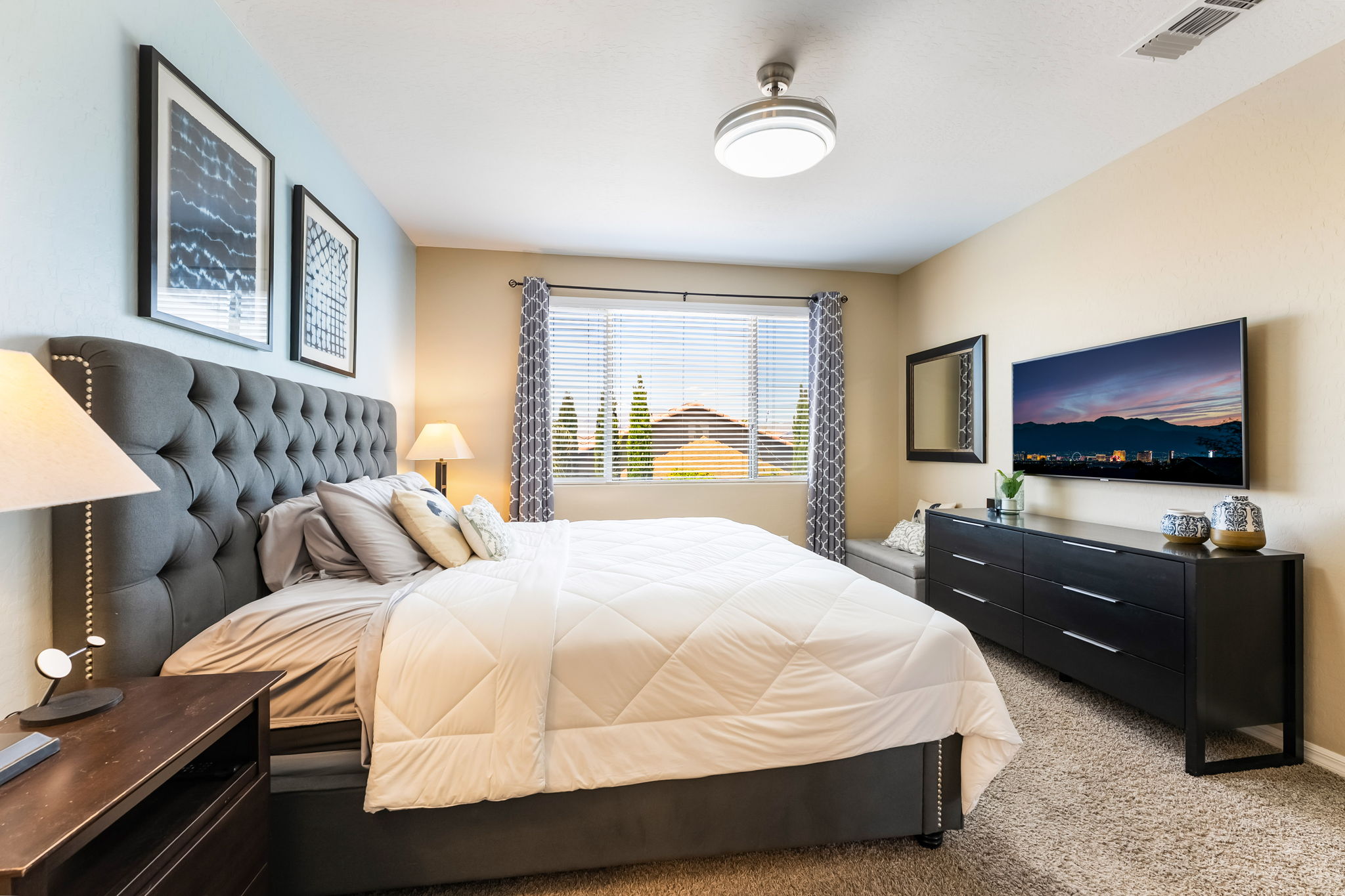 This primary bedroom features a large, tufted grey upholstered headboard as the focal point, complemented by a white quilted bedding set. A sleek black dresser sits against the wall beneath a wall-mounted television, while a window with patterned curtains allows natural light to fill the room. The space is finished with neutral wall tones and carpeted flooring, creating a comfortable and inviting atmosphere.