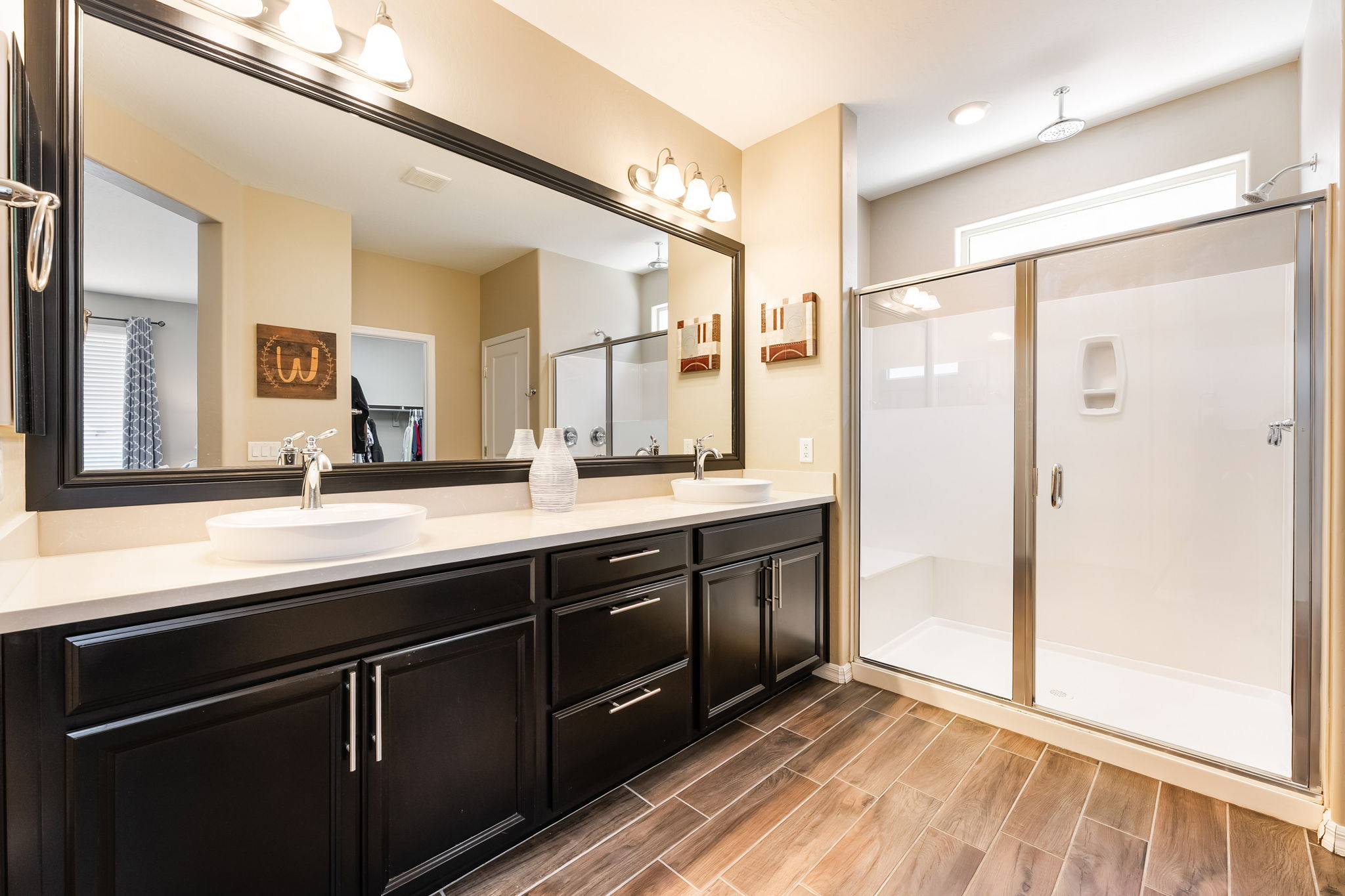 This primary bathroom features a long, dark-toned double vanity with a light-colored countertop and two vessel sinks. A large, framed mirror spans the length of the vanity, reflecting the adjacent walk-in shower with its glass enclosure and neutral-toned walls. The space is finished with wood-look tile flooring, creating a warm and cohesive aesthetic.