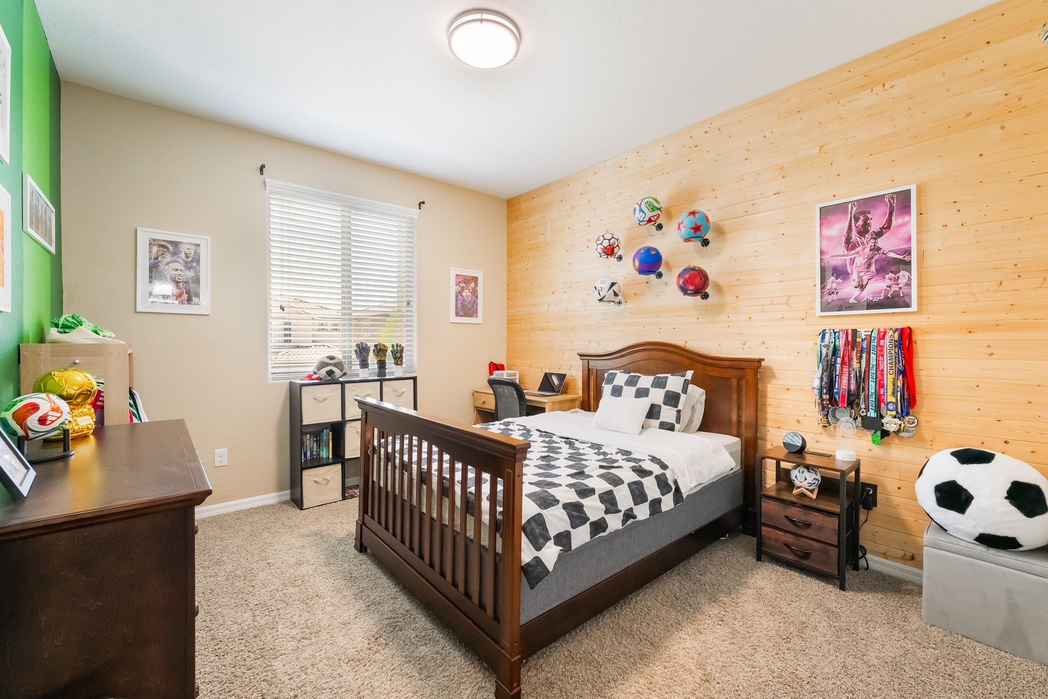 This themed bedroom features a prominent wood-paneled accent wall adorned with soccer balls and sports memorabilia, creating a personalized space for a sports enthusiast. The room includes a dark wood bed frame with a checkered comforter, a matching nightstand, and a storage unit with fabric bins. A large window with blinds allows natural light to brighten the room, which is finished with neutral carpeting and a green accent wall.
