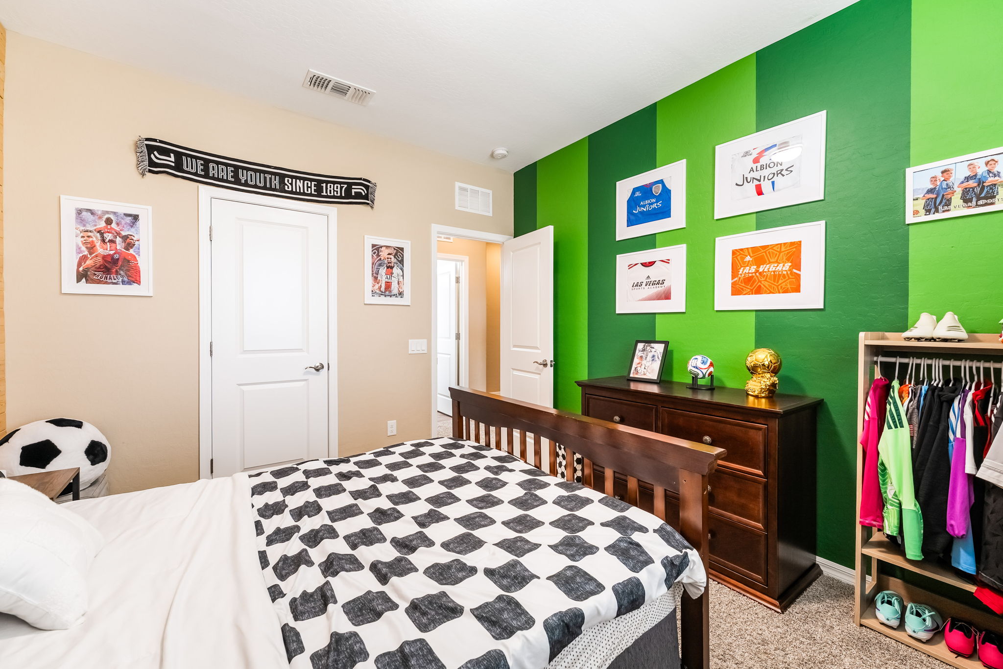 This youth-themed bedroom features a soccer-inspired decor with a green-striped accent wall adorned with framed jerseys and sports memorabilia. The room includes a wooden bed frame with a black-and-white checkered comforter, a dark wood dresser, and an open-concept clothing rack. The space is bright and functional, showcasing a personalized aesthetic suitable for a child or teenager.