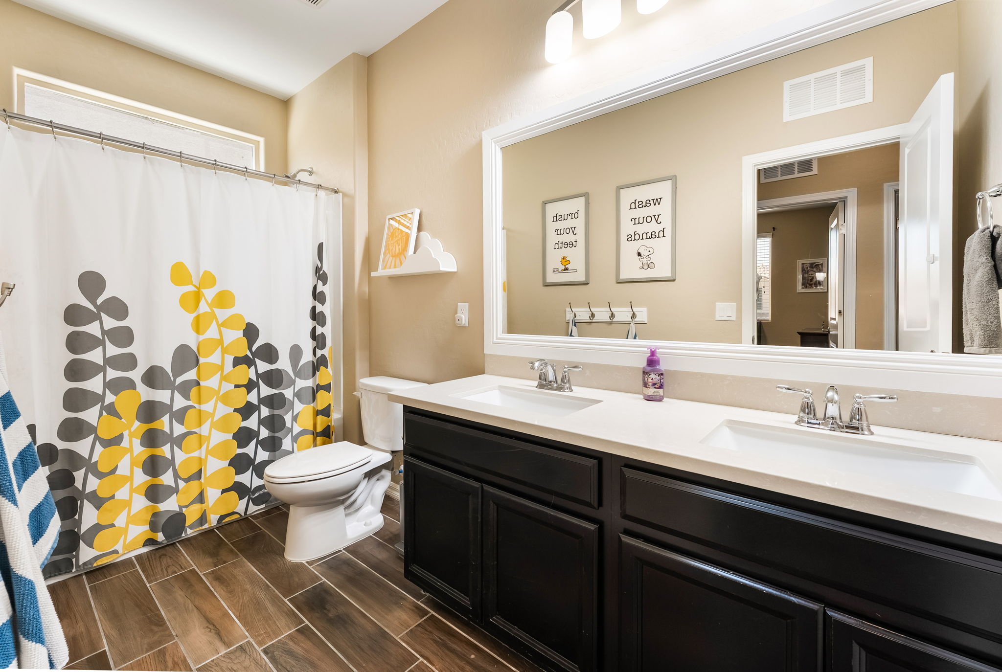 This bright and playful bathroom features a double vanity with dark cabinetry topped by a light-colored counter and dual sinks. A large, framed mirror spans the wall above the vanity, reflecting the shower area which is enclosed by a patterned shower curtain featuring yellow and gray leaf motifs. The space is finished with wood-look tile flooring and whimsical wall art, creating a cheerful and functional atmosphere.