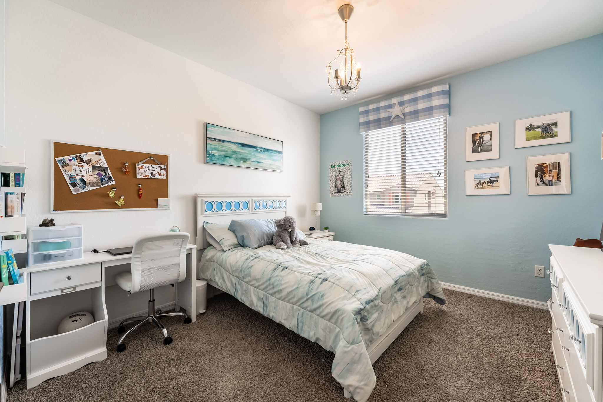 This bright and inviting bedroom features a light blue accent wall and a white bed frame with a patterned blue and white comforter. A dedicated study area with a white desk and rolling chair is positioned to the left, while a dresser sits on the right side of the room. The space is well-lit by a central chandelier and a window dressed with a blue and white checkered valance, creating a cozy and youthful atmosphere.