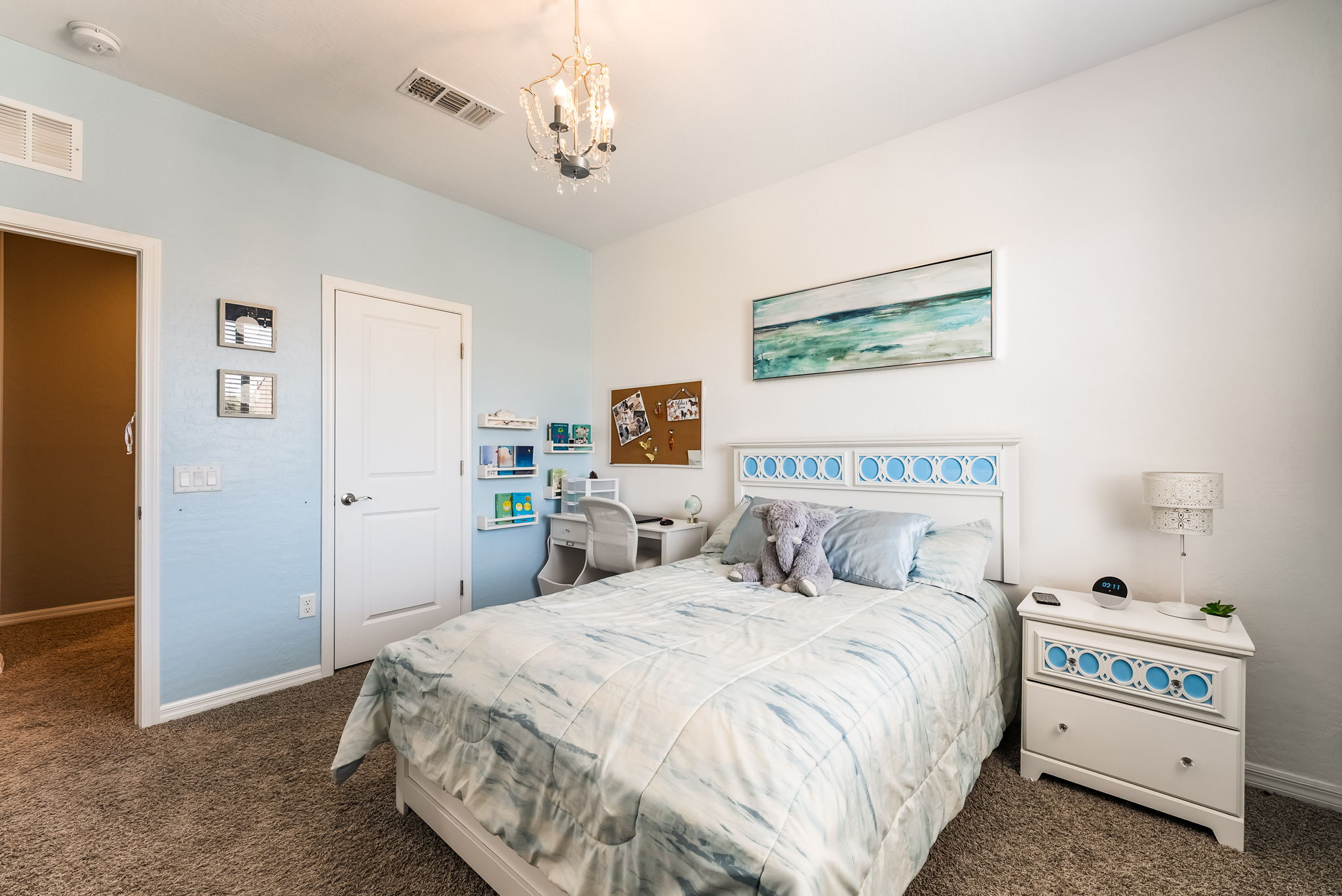 This bright and inviting bedroom features a light blue accent wall and a white bed frame with a matching nightstand. A soft, watercolor-inspired bedding set complements the room's coastal aesthetic, while a small desk area and a crystal chandelier add functional and decorative charm. The room is well-lit and organized, creating a cozy and serene atmosphere suitable for a guest or child's bedroom.