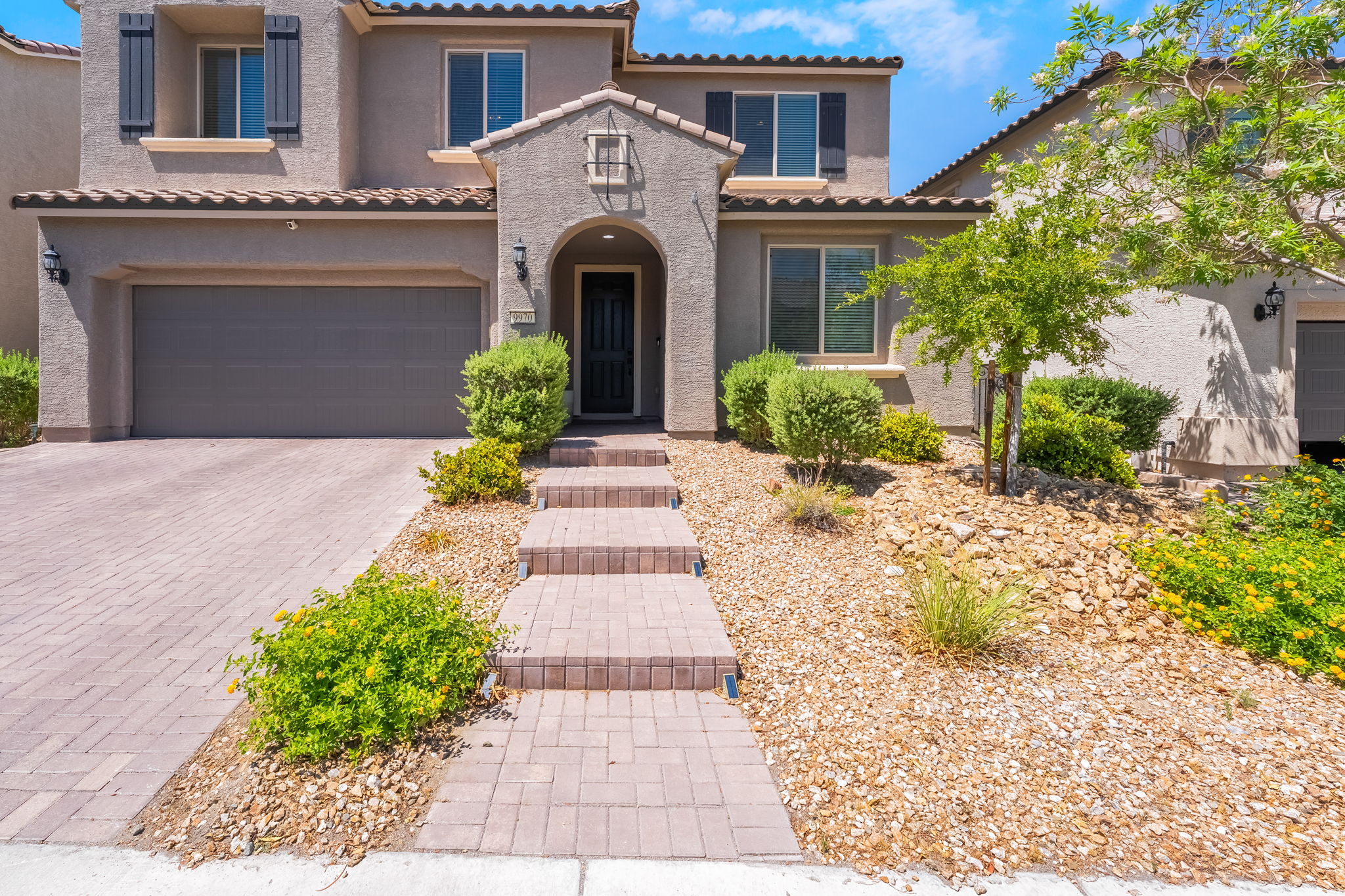 This two-story suburban home features a stucco facade with a neutral earth-tone finish and a tiled roof. A paved walkway with integrated step lighting leads to the arched entryway, complemented by a landscaped front yard with desert-style gravel and mature shrubs. The property includes an attached garage and a large window on the upper level, presenting a well-maintained and inviting curb appeal.