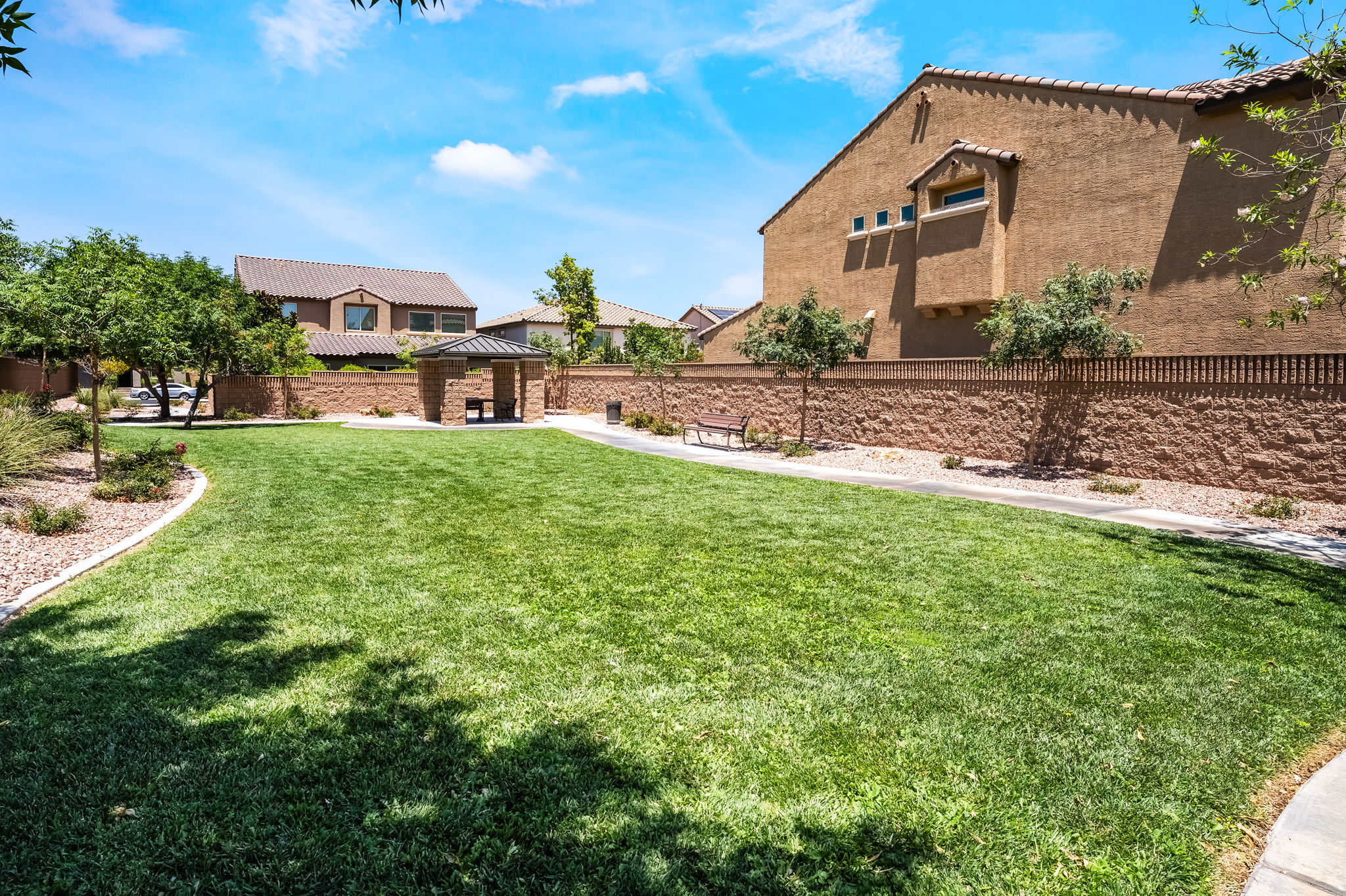 This community park area features a well-manicured green lawn bordered by a concrete walking path and a sturdy masonry wall. A covered gazebo with a picnic table provides a shaded seating area, complemented by a nearby park bench under the trees. The scene is captured from a ground-level perspective, showcasing a bright, sunny day in a residential neighborhood setting.