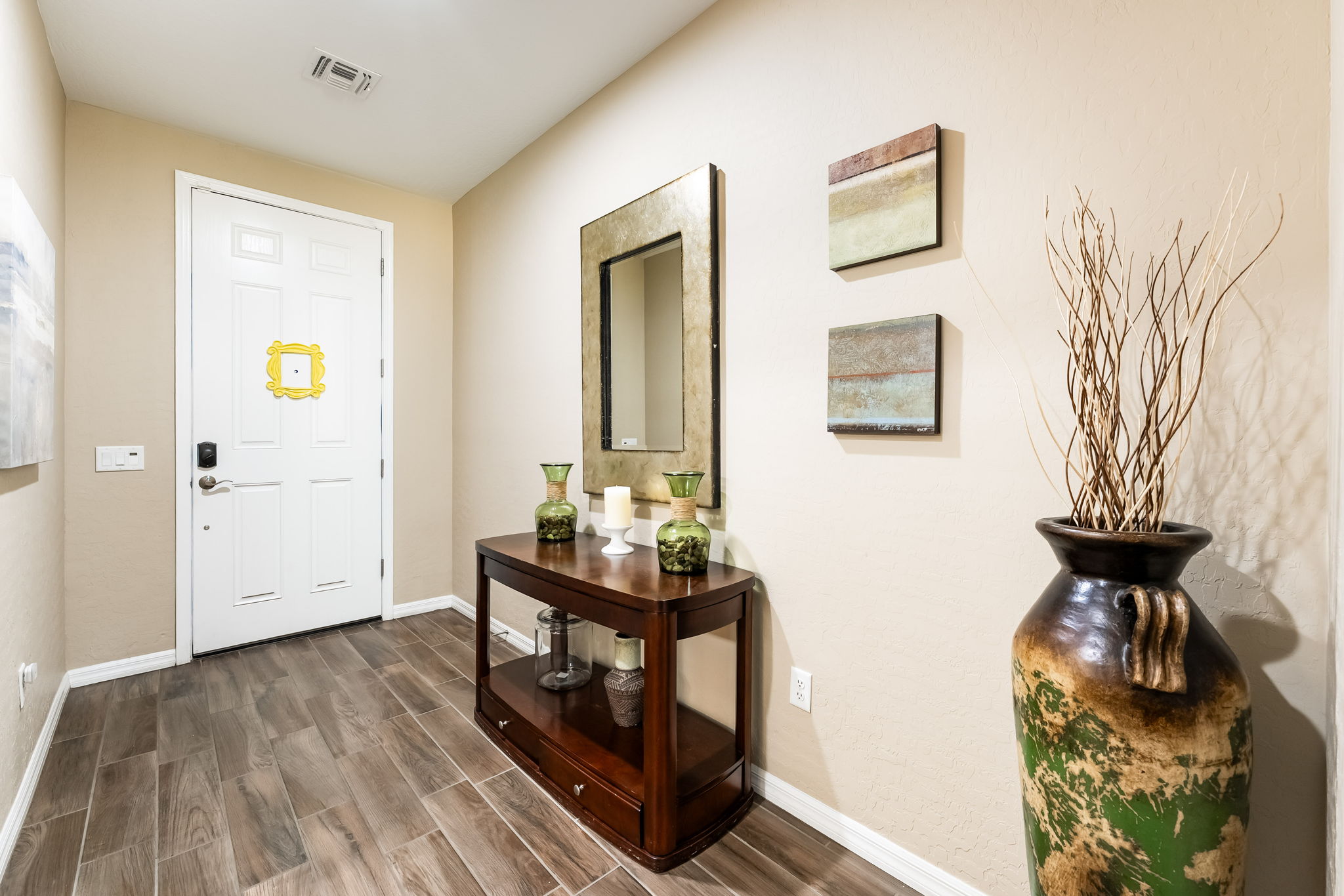 This welcoming entryway features wood-look tile flooring and a neutral color palette, creating a warm and inviting atmosphere. A dark wood console table sits against the wall, topped with decorative glass vases and a candle, while a rectangular mirror hangs above it. The space is accented by two pieces of abstract wall art and a large, rustic floor vase filled with decorative branches, providing a balanced and polished aesthetic.