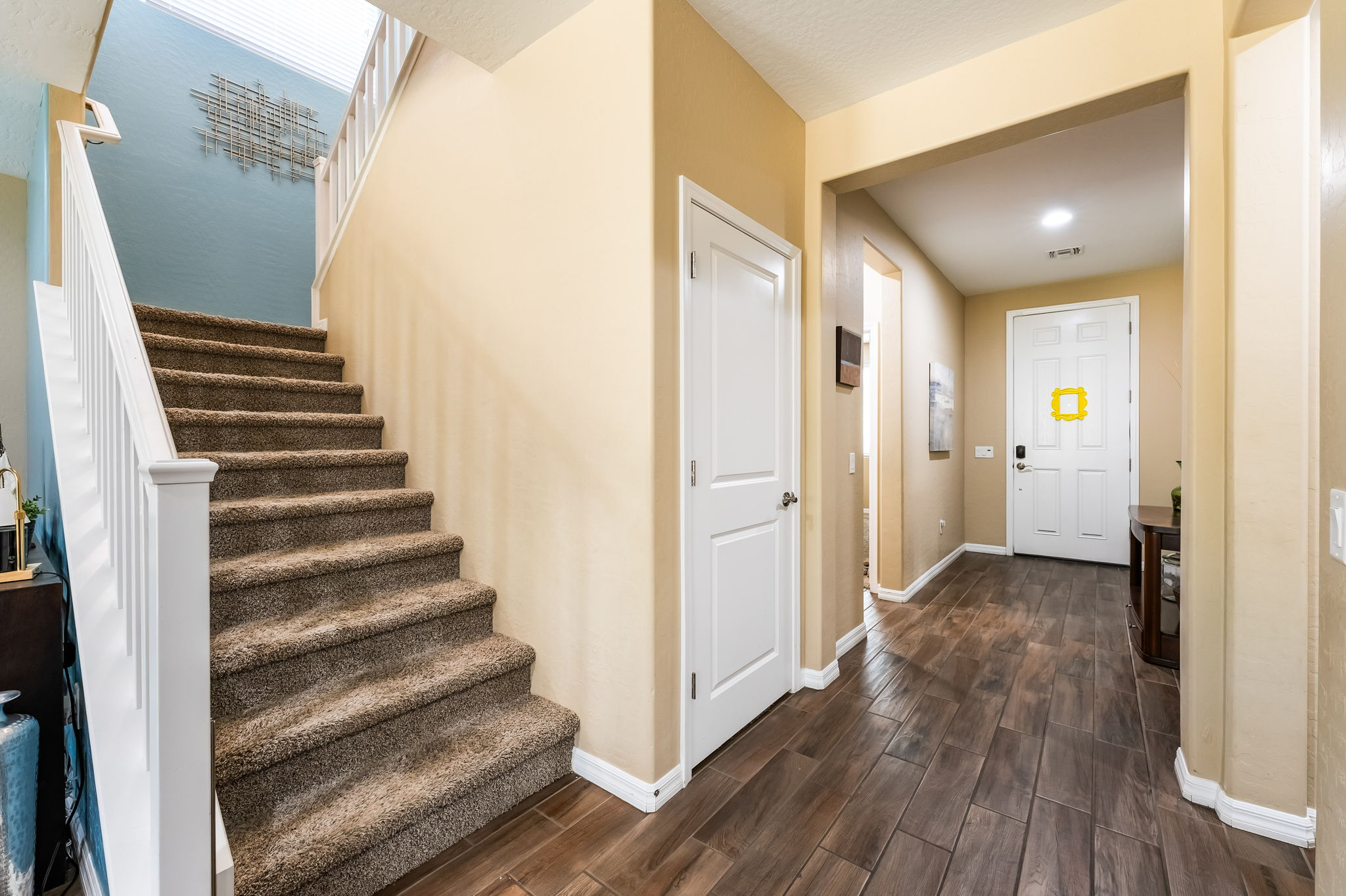 This entryway features a staircase with carpeted treads and a white railing, set against a light blue accent wall with a decorative metal wall sculpture. The hallway leads toward the front door, which is adorned with a yellow frame, and is finished with dark wood-look flooring and neutral-toned walls. The perspective captures the transition from the staircase area into the main entrance, highlighting the home's welcoming and well-maintained layout.