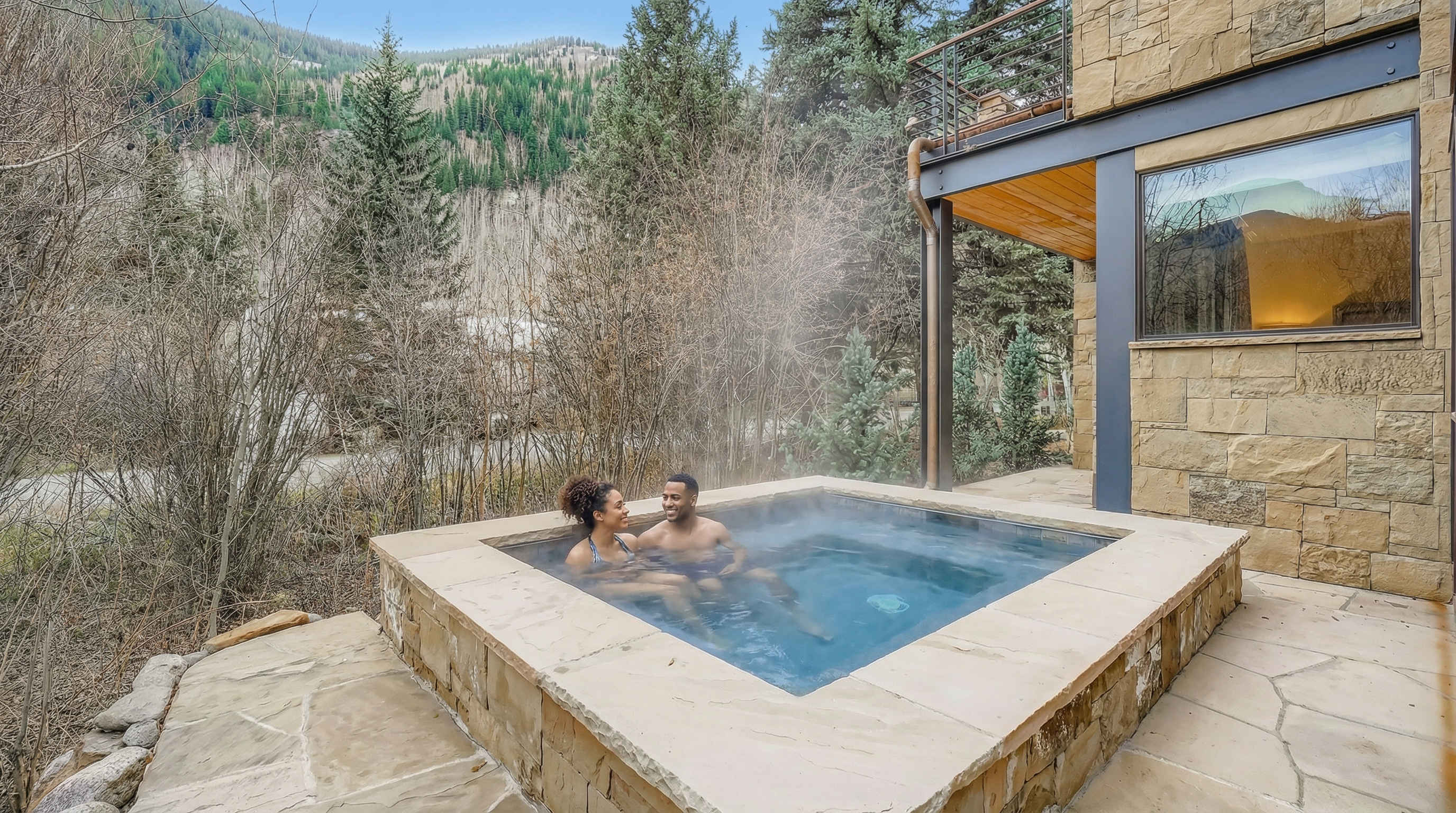 The image showcases an outdoor spa area with a couple enjoying the hot tub. The spa is surrounded by stone construction and landscaping, blending into the natural environment of trees and a mountain backdrop. It presents a high-end, serene ambiance, emphasizing relaxation and privacy.