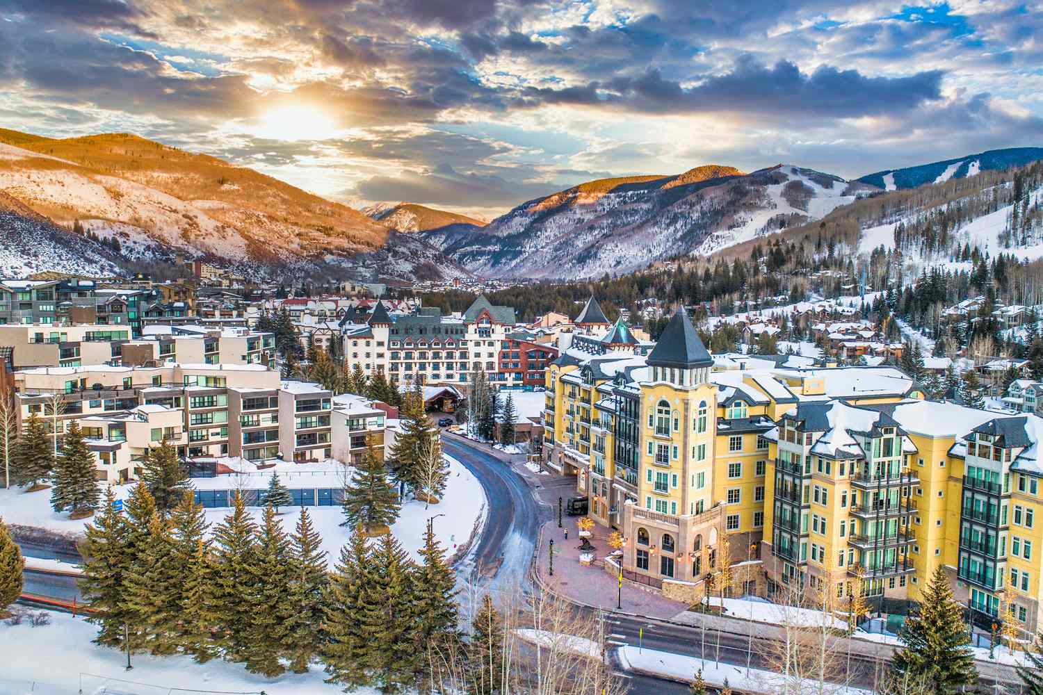 This aerial shot showcases a luxurious resort town nestled in a snowy mountain valley. The architecture features a blend of modern condos and European-inspired buildings, creating a charming and upscale atmosphere. With snow-covered peaks in the background, the stunning scenery adds to the appeal and offers potential for winter recreation.