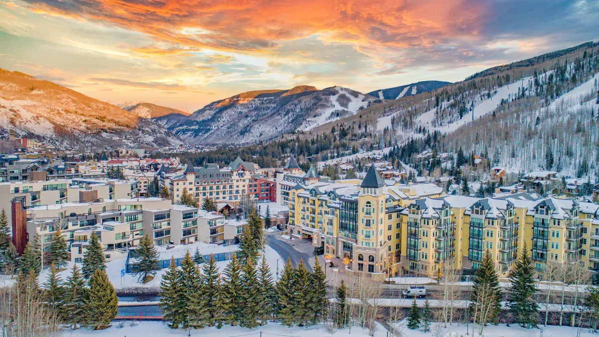 This aerial shot showcases a luxurious mountain resort town at sunset. Snow-capped mountains surround the town, which features upscale hotels and condominiums with distinctive architectural details. The warm sunset colors complement the wintery landscape, creating an inviting and picturesque scene appealing to potential buyers seeking a premium alpine retreat.