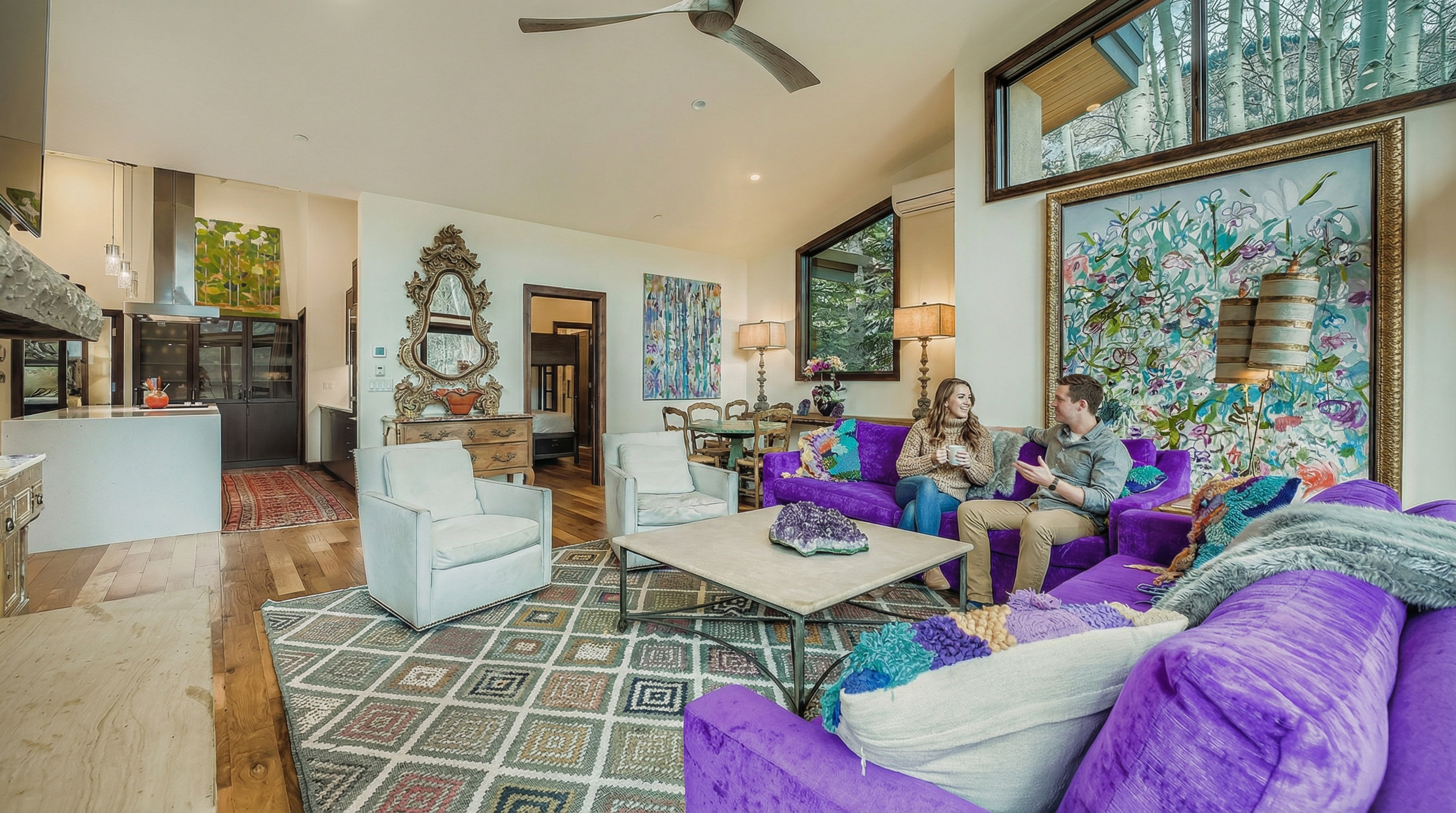 The living room is spacious and inviting, featuring a vibrant purple sofa and two white armchairs arranged around a geometric patterned rug and a coffee table. Artwork adorns the walls, enhancing the room's aesthetic appeal. A large window allows natural light to flood the space, and the open layout connects seamlessly with the kitchen, creating a welcoming atmosphere.
