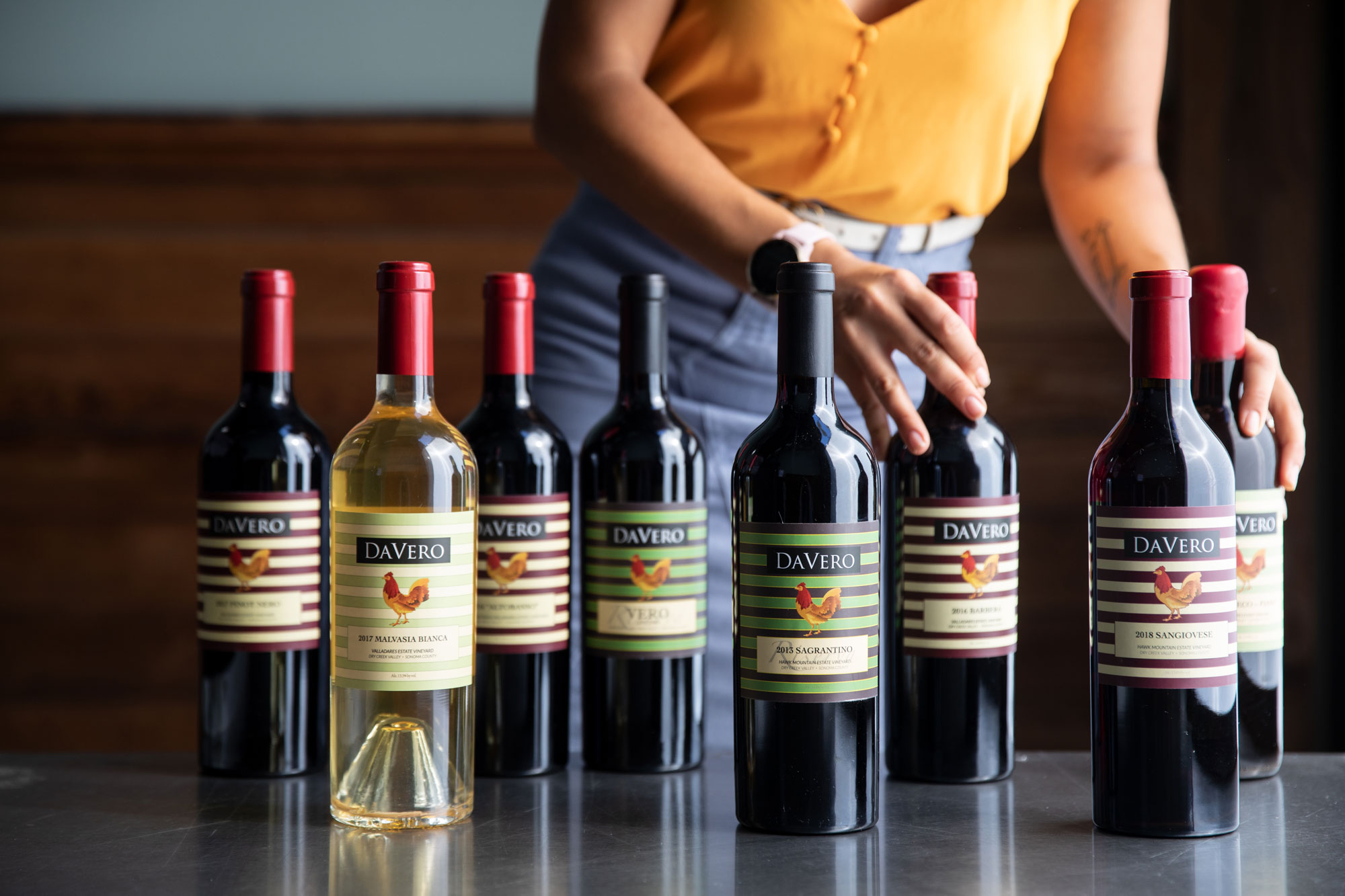 This image features a collection of DaVero wine bottles lined up on a stainless steel surface, with a person adjusting one of the bottles in the background. The arrangement highlights various vintage labels and consistent branding, suggesting a professional tasting or cellar setting. The warm lighting and wooden background provide a sophisticated, elegant aesthetic suitable for a winery or culinary feature.