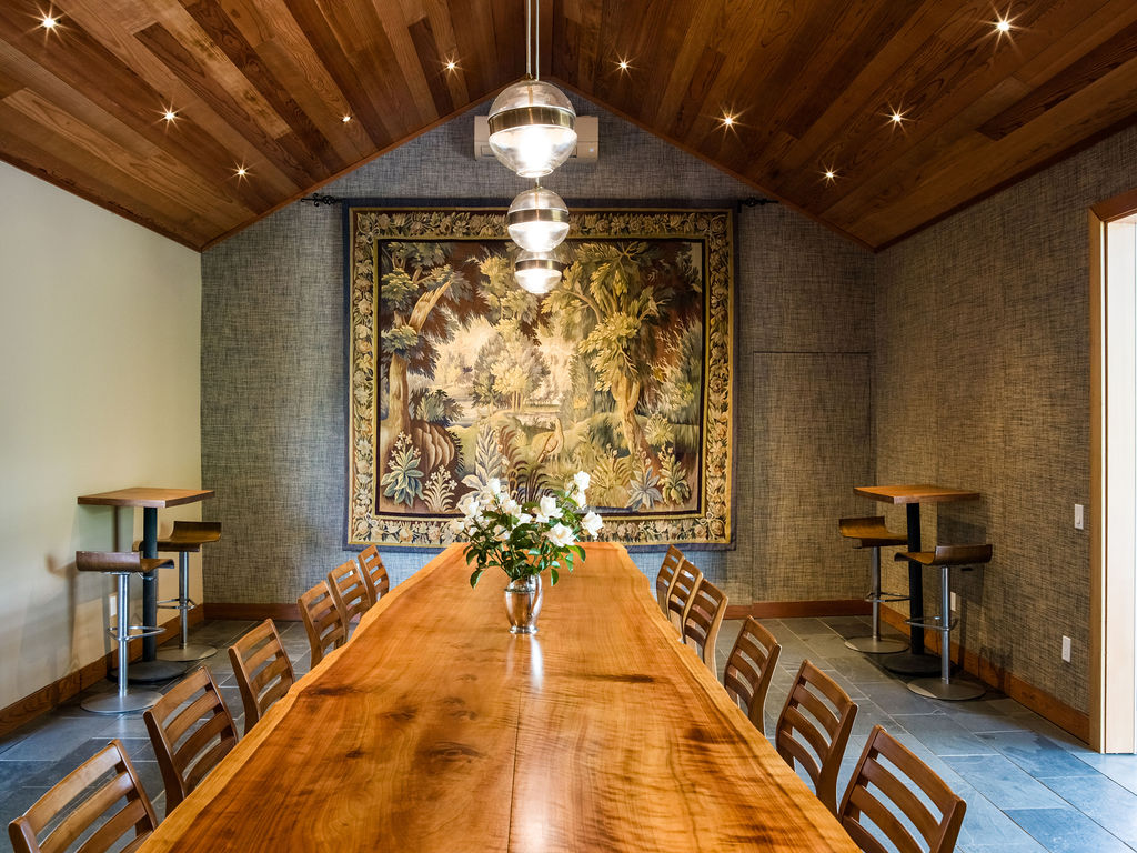 This spacious formal dining area features a striking long wooden slab dining table as the centerpiece, surrounded by modern wooden chairs. The room boasts a vaulted wood-paneled ceiling with recessed lighting and a sophisticated tapestry hanging on the back wall, complemented by elegant pendant fixtures. The combination of high-end materials and warm lighting creates a refined and inviting atmosphere.