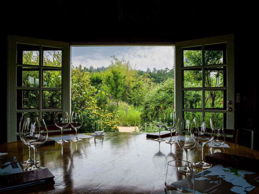 This interior dining space features a large, polished wooden table set with multiple wine glasses, suggesting a dedicated tasting or gathering area. The room is anchored by a wide, open window frame that provides a stunning, unobstructed view of a lush green garden. The combination of natural light and serene garden views creates an inviting, upscale atmosphere perfect for social dining.